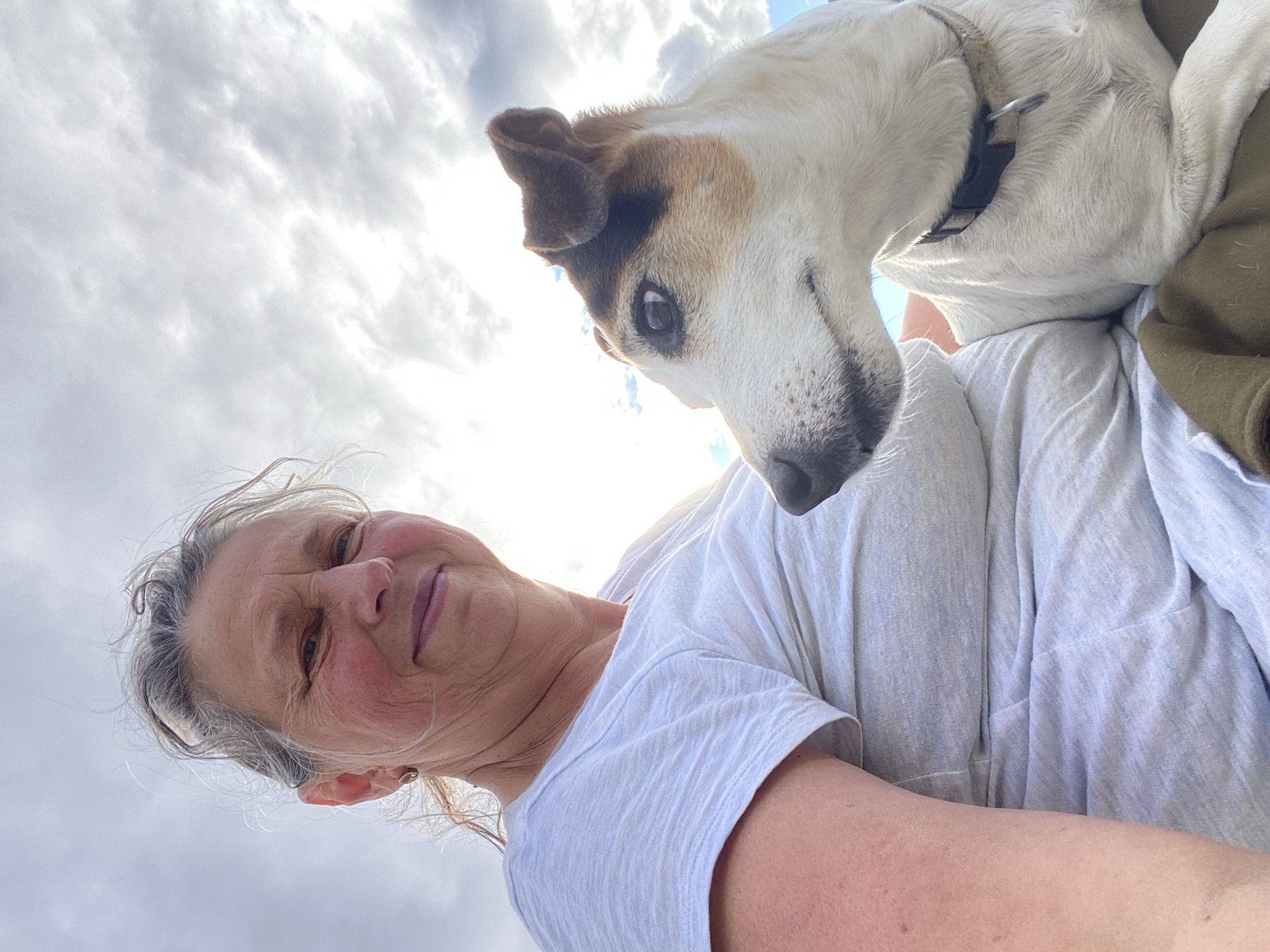 A woman with grey hair pulled back and a white t-shirt smiles at the camera, her face partially obscured by the bright sky. To her right, a small white and brown dog with its head turned to the side, wearing a dark collar, rests its head on her shoulder. The background is a bright, cloudy sky with the sun shining through. The image appears to have been taken outdoors, possibly in a park or natural setting. The location in the city of Yukon, Unorganized, Canada is not evident from the image.