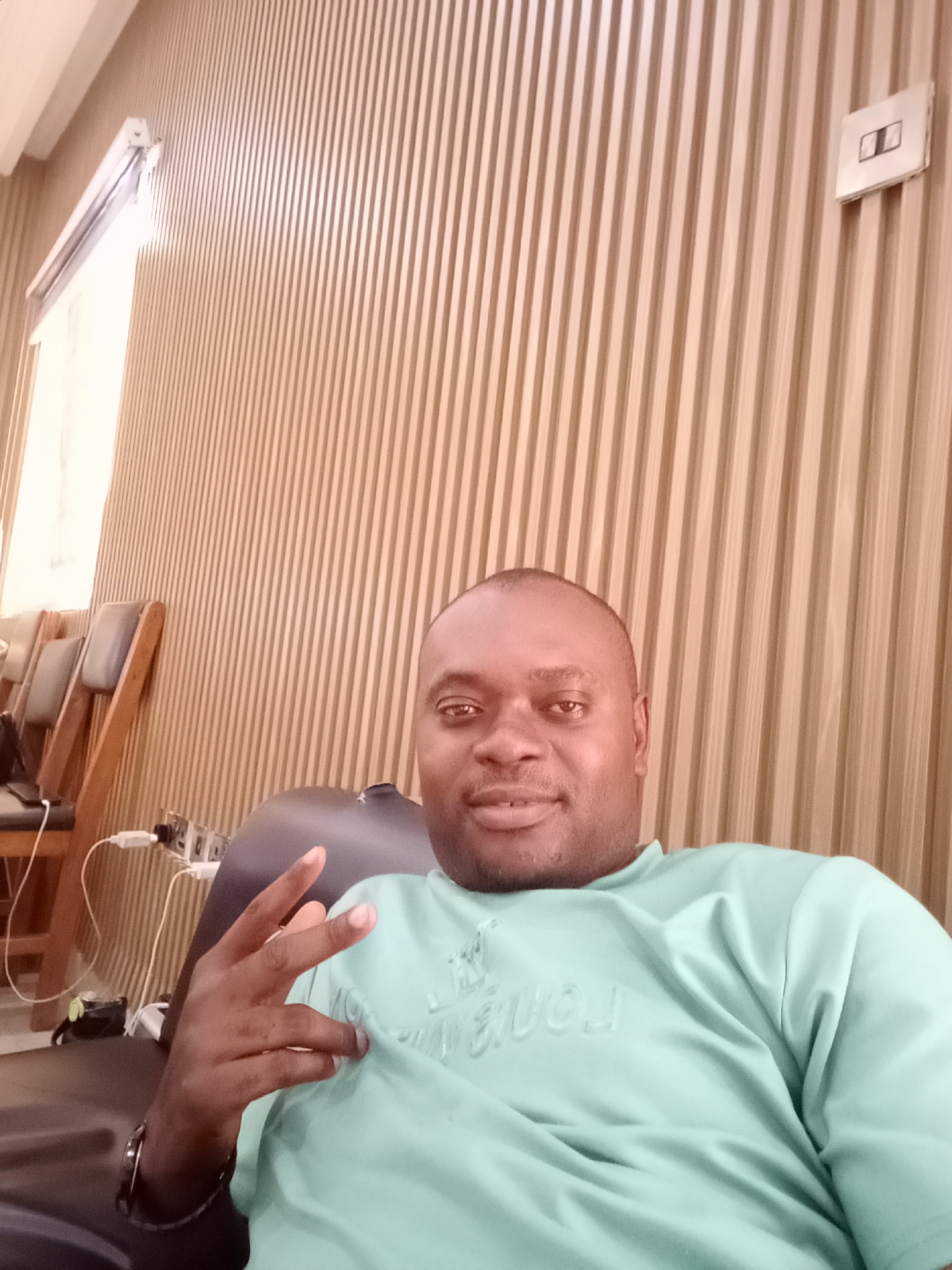 A medium shot depicts a man in a light green t-shirt, sitting indoors and giving a peace sign. The background features vertical wood paneling with a window on the left side and a light switch on the right. Wooden chairs with grey cushions are partially visible to the left of the man. Power adapters and chargers are visible, suggesting a place of rest. Based on the prompt's context, the location is Gwarinpa, Nigeria.