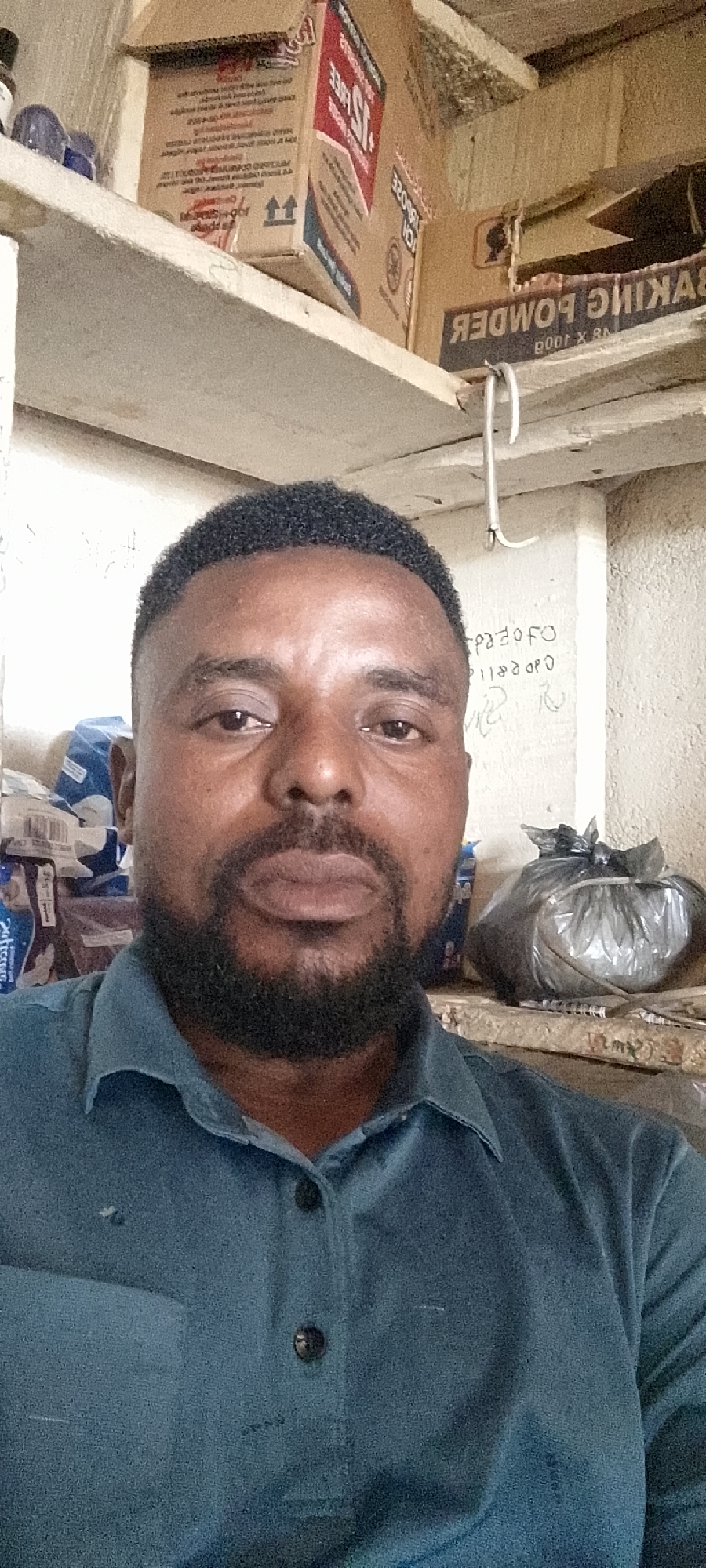 This is a selfie taken by a Black man with dark skin, short black hair, and a beard. He is wearing a teal polo shirt. Behind him, shelves are visible. On the top shelf are several cardboard boxes, one with the text "BAKING POWDER" visible and partially reversed. On the middle shelf, a plastic-wrapped bundle is visible to the right. The man is positioned in the foreground, with the shelves and their contents in the background. The scene appears to be indoors, possibly a shop or storage area. The location is identified as Durumi, Nigeria.
