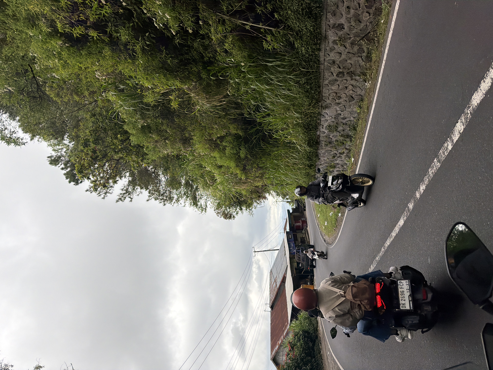 The image depicts a road scene in Kintamani, Indonesia, under an overcast sky. In the foreground, two people are riding on a scooter, with the driver wearing a red helmet and a beige jacket, and the passenger wearing a backpack. The scooter has a license plate that reads "DK 2696 FCP". Further down the winding asphalt road, another scooter is visible with a person wearing a gray jacket and helmet. To the right of the road, a lush green hillside is covered in dense vegetation and trees, with a stone wall lining the roadside. On the left side of the road, a few buildings with rusted metal roofs and some smaller structures are present, alongside more greenery. Power lines are strung across the sky above the buildings. The overall atmosphere is one of everyday travel on a scenic route.