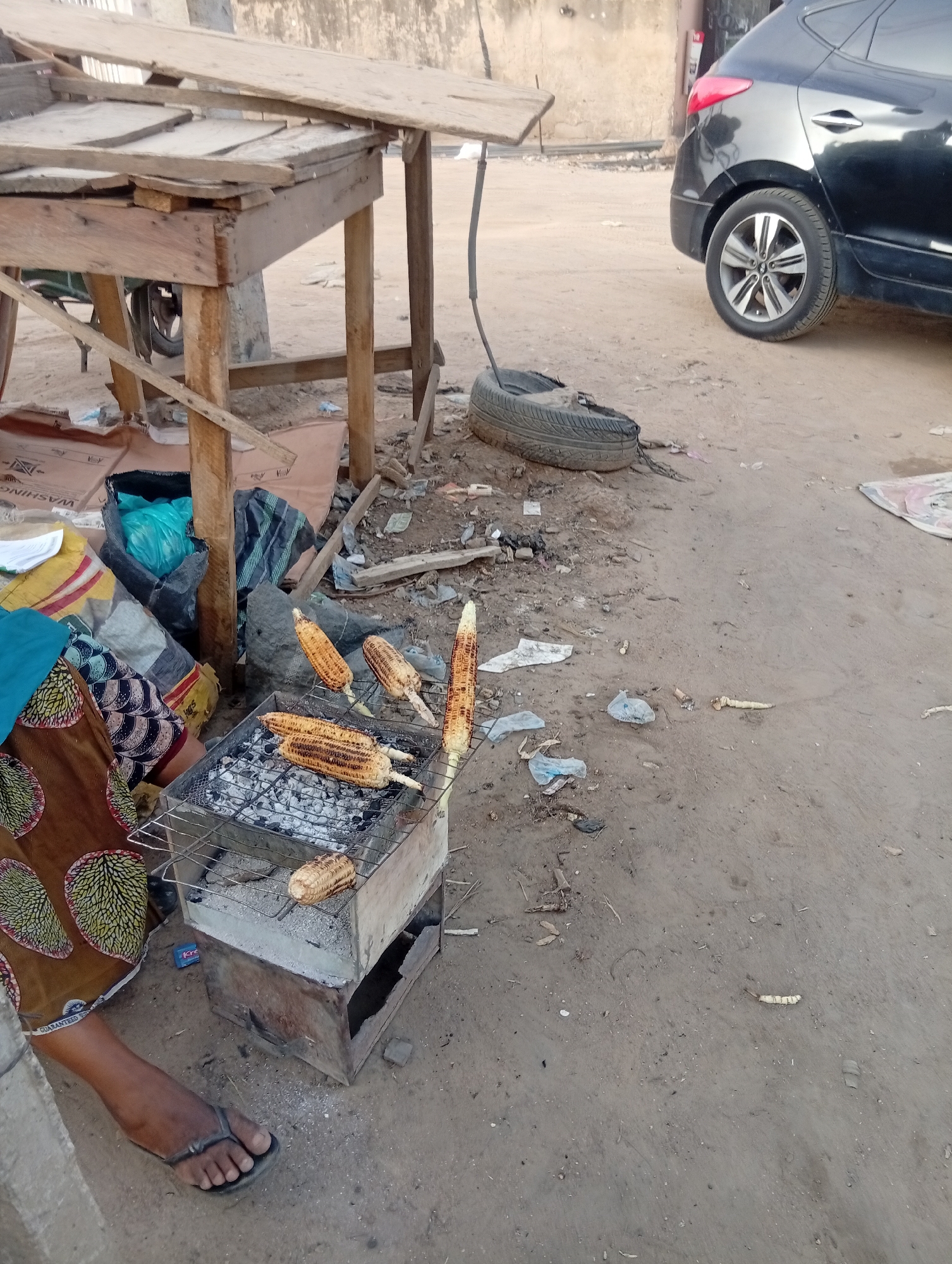The image depicts an outdoor scene in Aviation Quarters, Nigeria. A person wearing a patterned brown skirt and black flip-flops sits next to a small charcoal grill, where several ears of corn are being roasted. The grill is made of metal and sits on a simple box-like structure. To the left of the grill is a pile of sacks and bags, and a makeshift wooden table stands nearby. The ground is littered with trash and debris. In the background, a black SUV is partially visible, parked on the dirt road. The environment appears to be a roadside setting, possibly a vendor's stall.