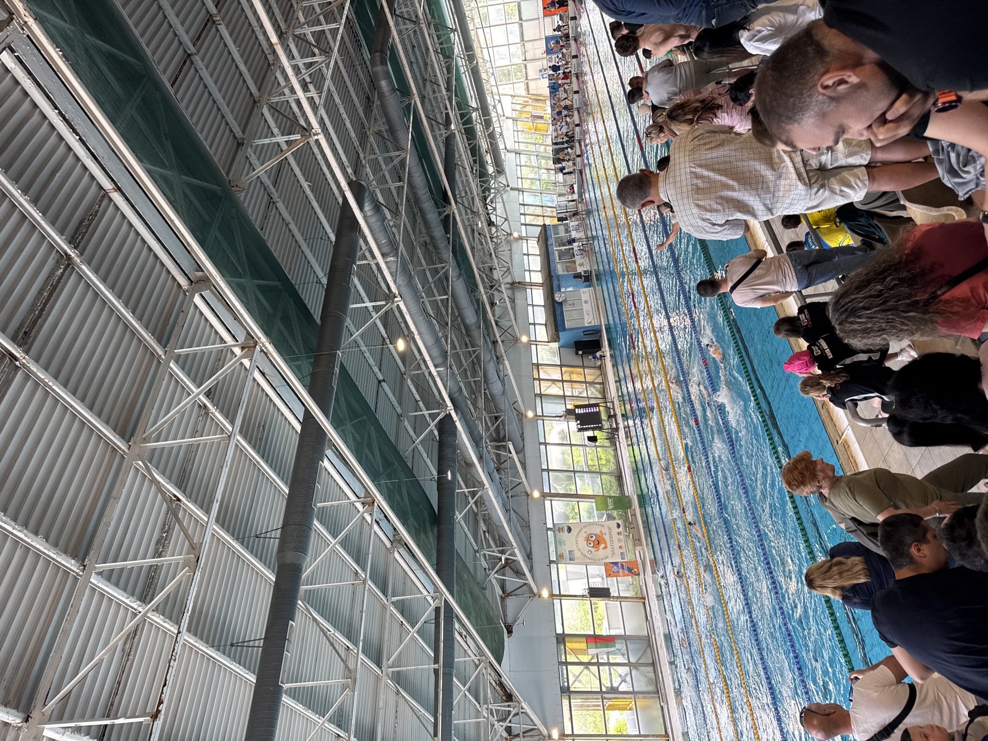 The image shows an indoor swimming pool, likely in Sofia, Bulgaria, based on the Cyrillic text visible in the image. The pool has multiple lanes with lane markers visible. Several people are standing or sitting along the edge of the pool, appearing to be spectators watching a swimming event. Some of the spectators are leaning over the edge of the pool, watching the swimmers. The indoor structure has a metal roof with pipes and ventilation. Light enters the pool area through windows along one side. A digital display board is visible near the windows, possibly showing race times or results. Banners or signage with text in Bulgarian are also visible near the windows. Swimmers are in the pool, participating in a race or swim practice.