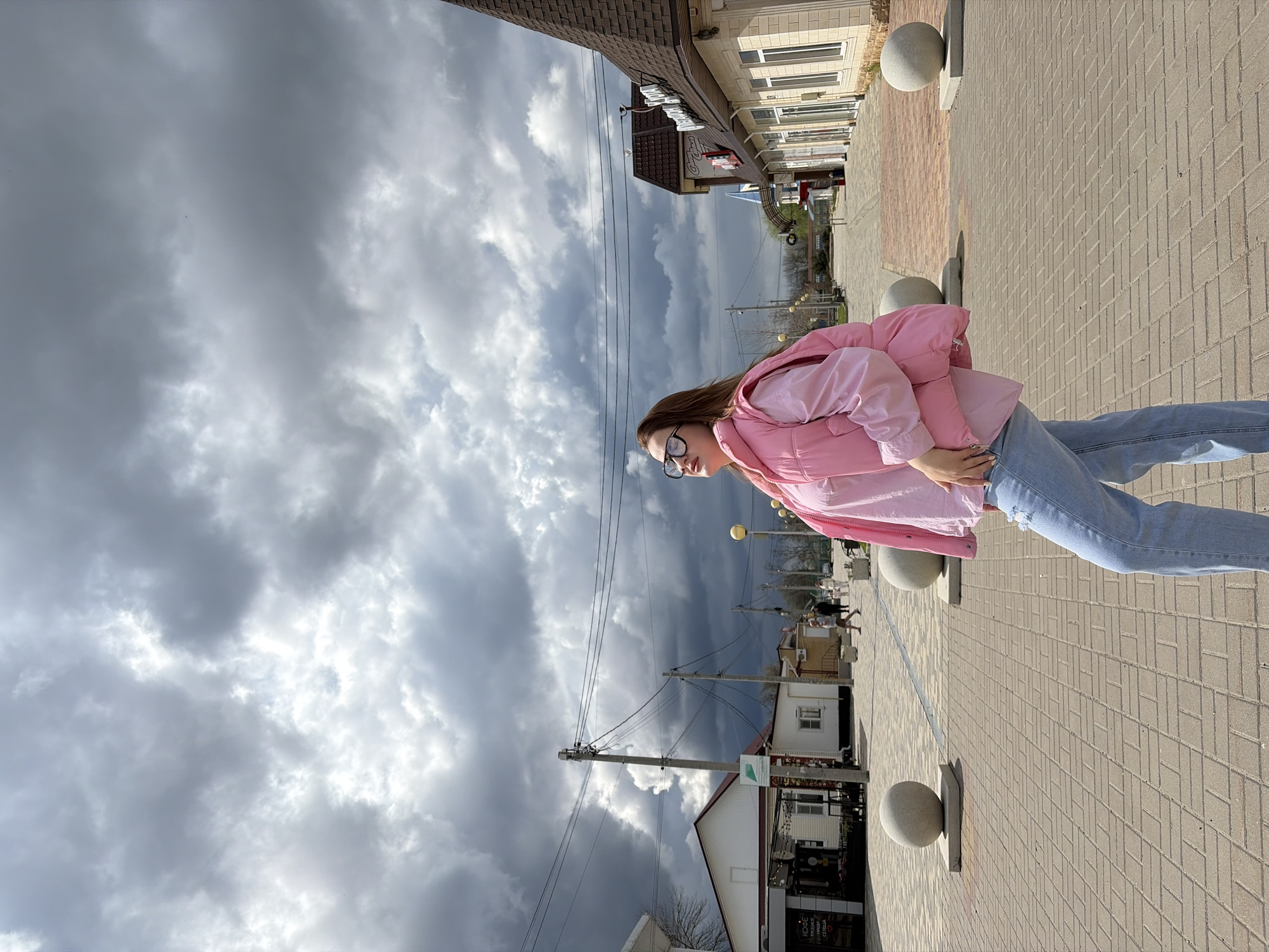 The image shows a young woman standing on a brick sidewalk in the city of Novopokrovskaya, Russia. She is wearing a pink jacket, a light pink top, blue jeans, and glasses. The sky is overcast with dark, dramatic clouds. Buildings line the street, some with storefronts visible. Utility poles and wires are also visible against the sky. There are stone spheres installed into the sidewalk along the street.
