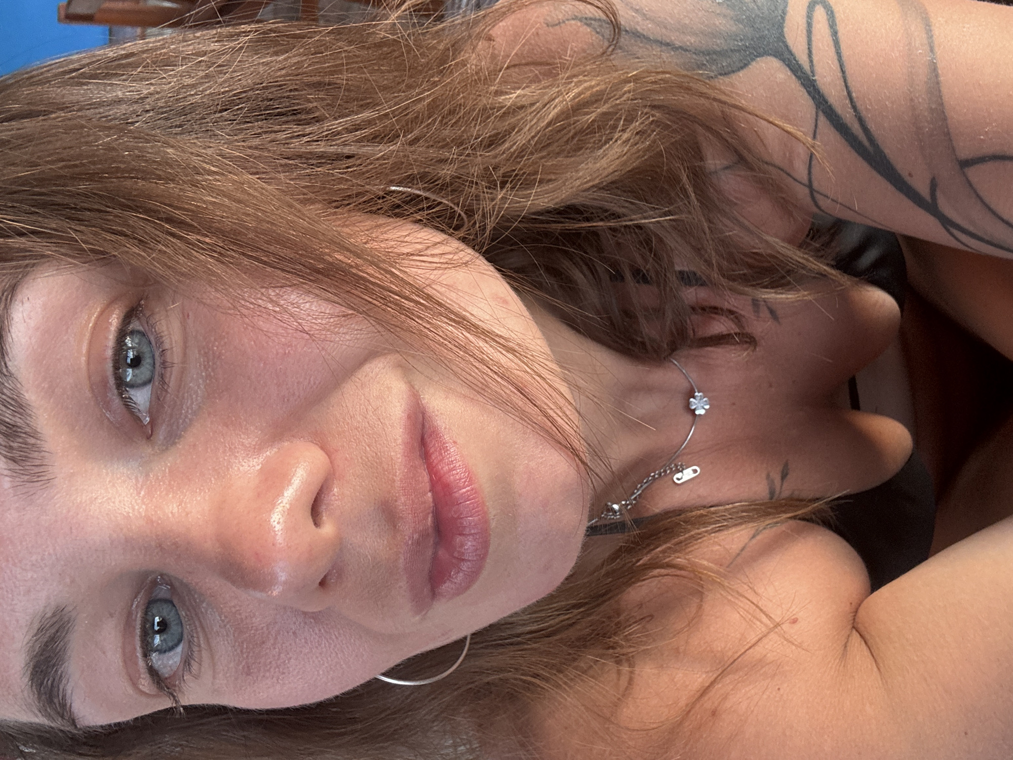 A close-up, angled portrait shows a woman with light brown hair and blue eyes. She has fair skin and is wearing a silver necklace and hoop earring. A tattoo is visible on her upper arm. The setting is unknown.