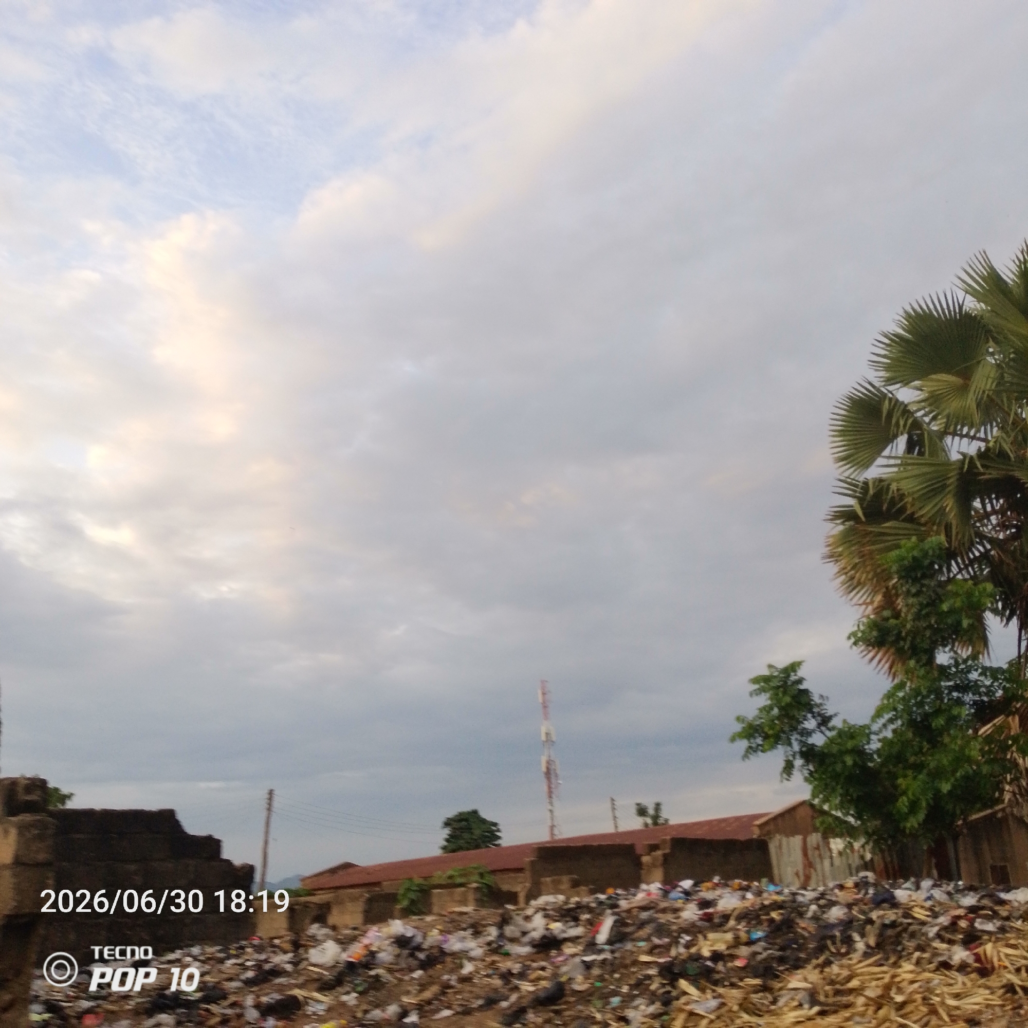 The media file depicts an outdoor scene with a large pile of refuse in the foreground. The trash consists of various items including plastic bags, wrappers, and what appear to be discarded wooden materials. In the background, there are several buildings with corrugated metal roofs and some brick structures. A tall telecommunications tower is visible in the distance. Lush green trees, including palm fronds, are scattered throughout the scene, some partially obscuring the buildings. The sky is overcast with dramatic clouds, suggesting a cloudy day, possibly with approaching rain. The overall impression of the scene is one of urban neglect and environmental concern due to the extensive garbage accumulation.
Visible text in the image includes a timestamp "2026/06/30 18:19" and a brand name "TECNO POP 10". The lighting suggests it is late afternoon or early evening. The location is identified as Jalingo, Nigeria. There are no people visible in the frame. The scene conveys a somber and potentially concerning atmosphere due to the waste management issues implied by the overflowing refuse.
