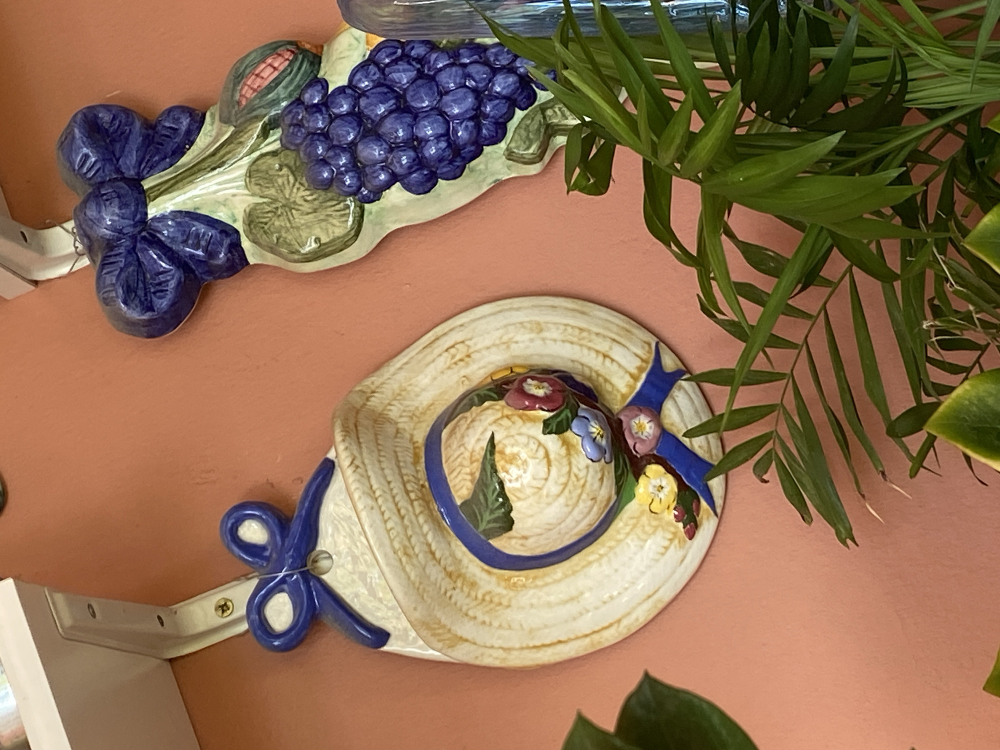 This media file depicts two ceramic wall decorations against a coral-colored wall. The upper decoration is a cluster of grapes with a cob of corn, rendered in shades of purple, green, and yellow. A bright blue bow is attached to the left side of this piece. Below it, a straw hat decoration features a blue ribbon adorned with small, colorful flowers. Blue bows are also attached to the brim and side of the hat. A metal shelf bracket is visible below and to the left of the hat. Green foliage from a houseplant hangs into the upper right portion of the frame, partially obscuring a glass object at the top. The lighting appears to be indoor, with no indication of time of day or weather. The overall scene suggests a home interior.
