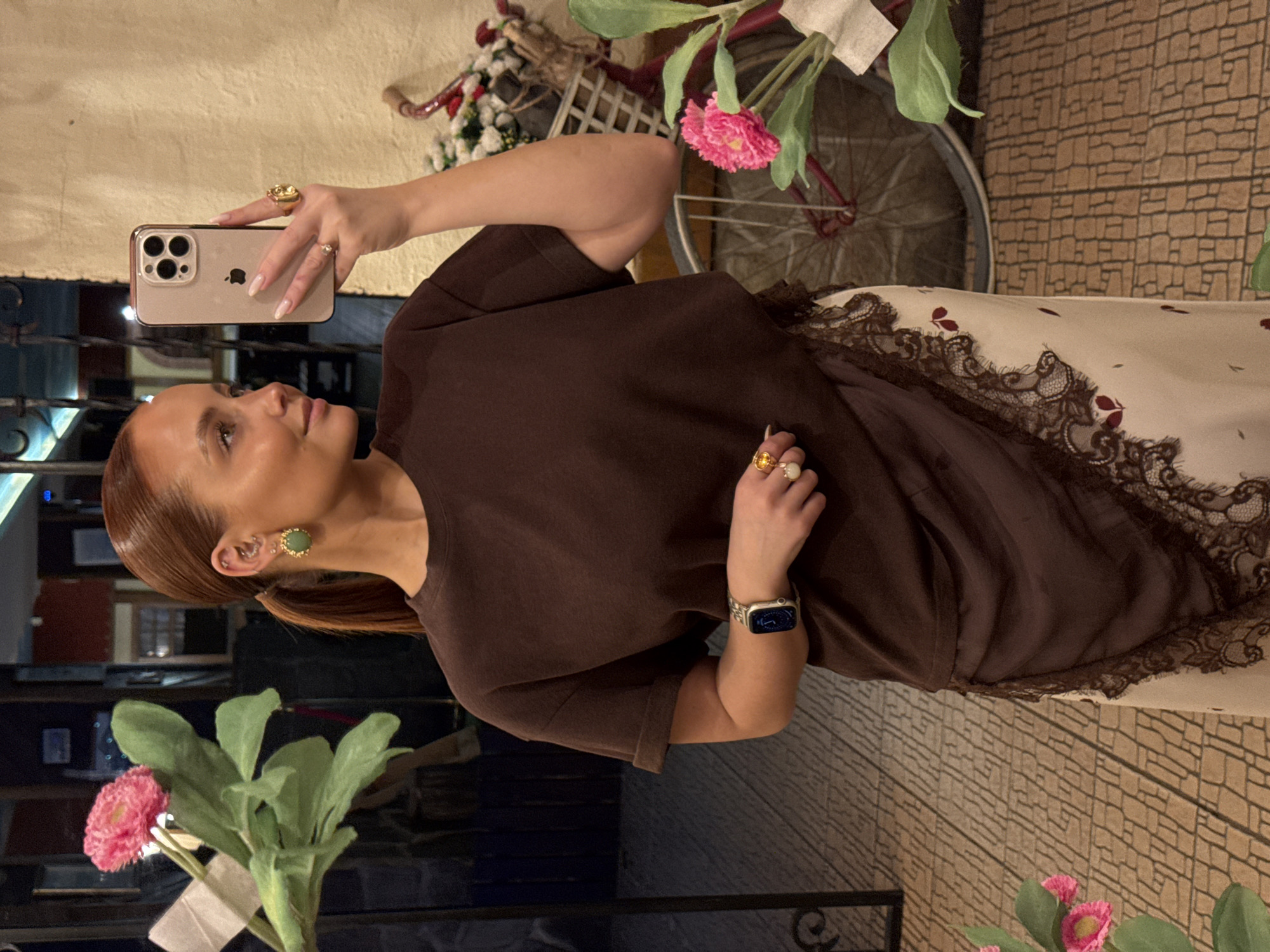 A woman with auburn hair pulled back in a ponytail is taking a selfie. She is wearing a brown t-shirt and a white skirt with a lace trim. She is wearing earrings, a ring on her left hand, and a watch on her left wrist. She is holding a phone in her right hand. In the background, there is a bicycle, flowers, and a brick wall. The location is unknown, but the request mentioned Chaikovsky, Russia.