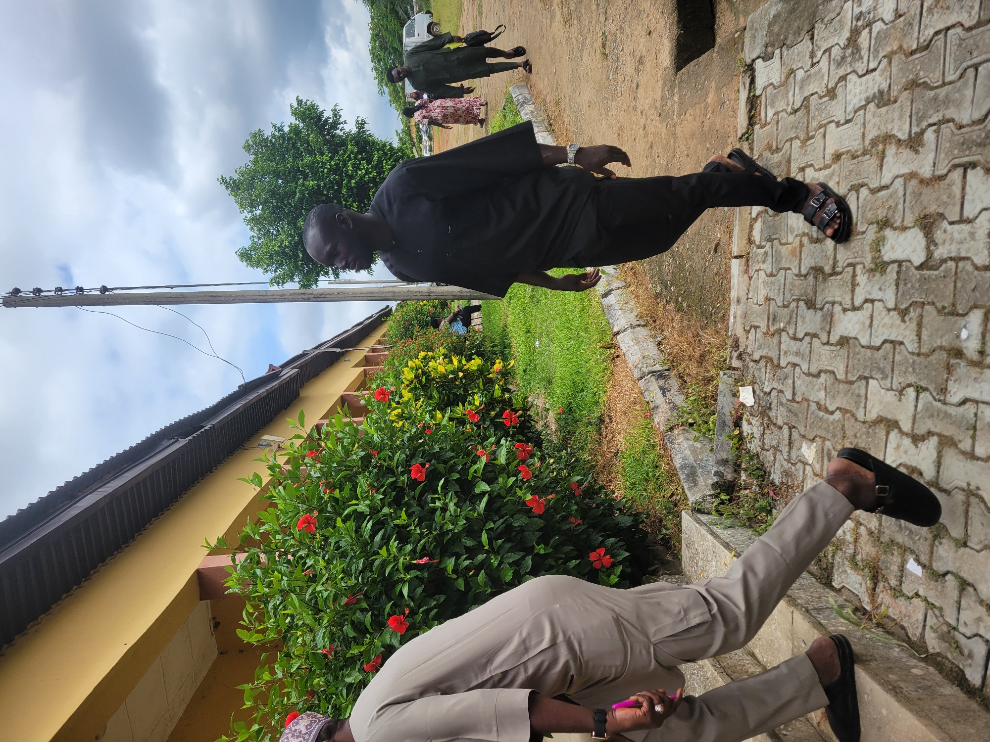 The image depicts an outdoor scene, likely on institutional grounds, within the city of Keffi, Nigeria. The sky is largely overcast with prominent grey clouds, though patches of blue are visible.
In the foreground left, a person is ascending concrete steps. This individual is primarily visible from the upper thigh down, wearing light brown traditional attire and black slip-on shoes. A black wristwatch is on the left wrist, and a pink object is held in the right hand. A portion of a patterned cap is visible at the top of the head.
In the mid-ground, a man with dark skin and short hair walks rightward on a paved path composed of interlocking concrete pavers. He wears a black traditional tunic with a buttoned placket, black trousers, and black double-strap sandals. A silver-colored wristwatch is on his left wrist.
To the left of the man, a single-story building with a yellow facade and a brown corrugated metal roof runs horizontally. In front of this building, a dense hedge of green bushes featuring numerous bright red hibiscus flowers extends along the path. Further along, a bush with variegated yellow-green leaves is visible.
Behind the man in black, a tall utility pole with wires extends upward. A large, dense green tree is situated behind the pole. In the far background right, two additional individuals are walking on an unpaved dirt path: one in a floral-patterned dress and another man in dark green traditional attire carrying a black bag. A white van or mini-bus is partially visible further in the background.
The ground surface consists of concrete pavers, dirt, and grass patches, delineated by concrete curbs. Small pieces of white litter are present on the paved areas.