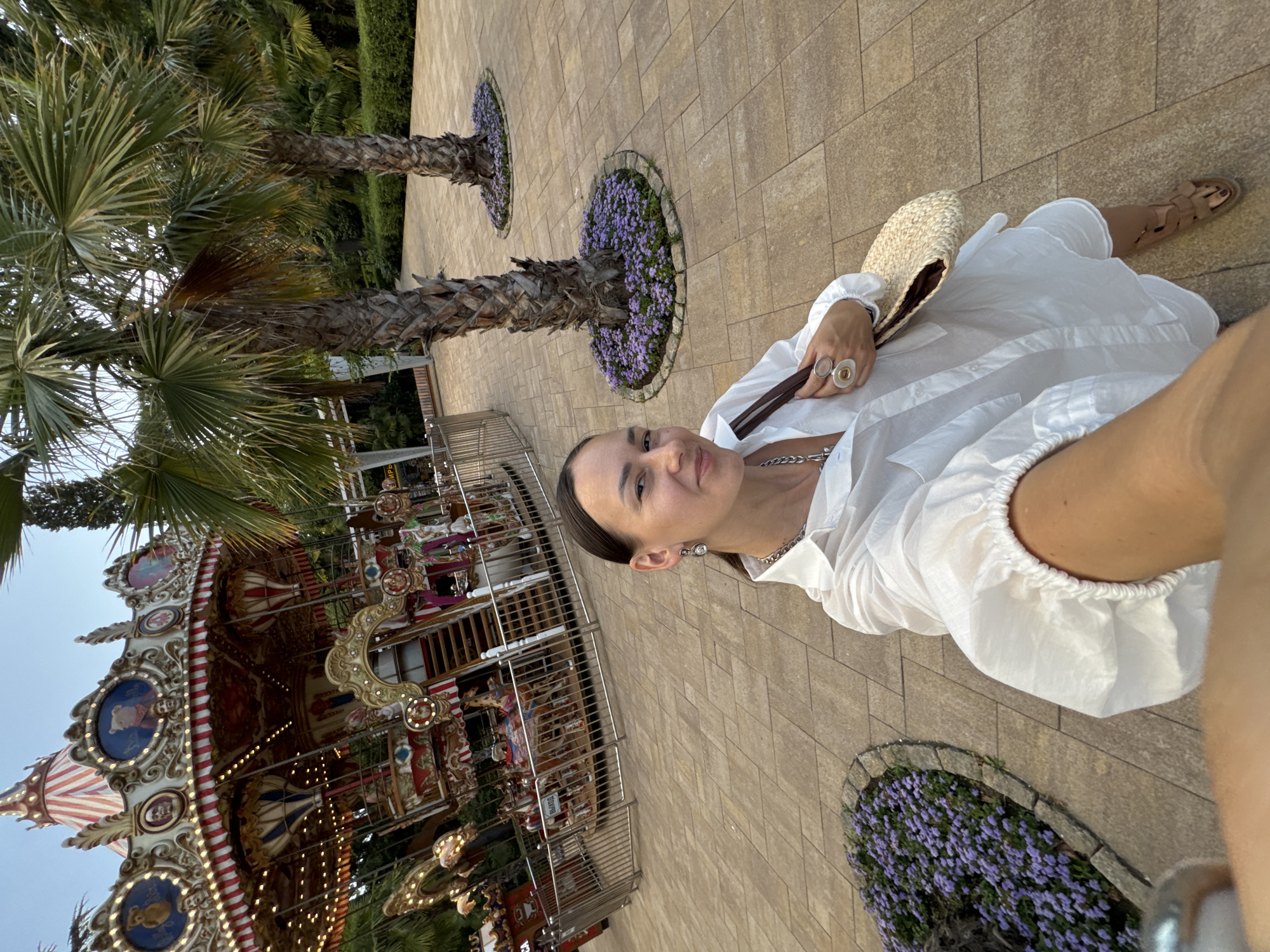 A woman is taking a selfie in a park with a carousel. She is wearing a white dress, sandals, and carrying a straw hat. She has two rings on her right hand. In the background, a carousel is visible, along with palm trees and purple flowers. The pavement is made of light brown tiles. The time of day appears to be late afternoon or early evening, as the light is soft. The weather seems pleasant. The scene evokes a relaxed and cheerful atmosphere. There is no visible text in the image.