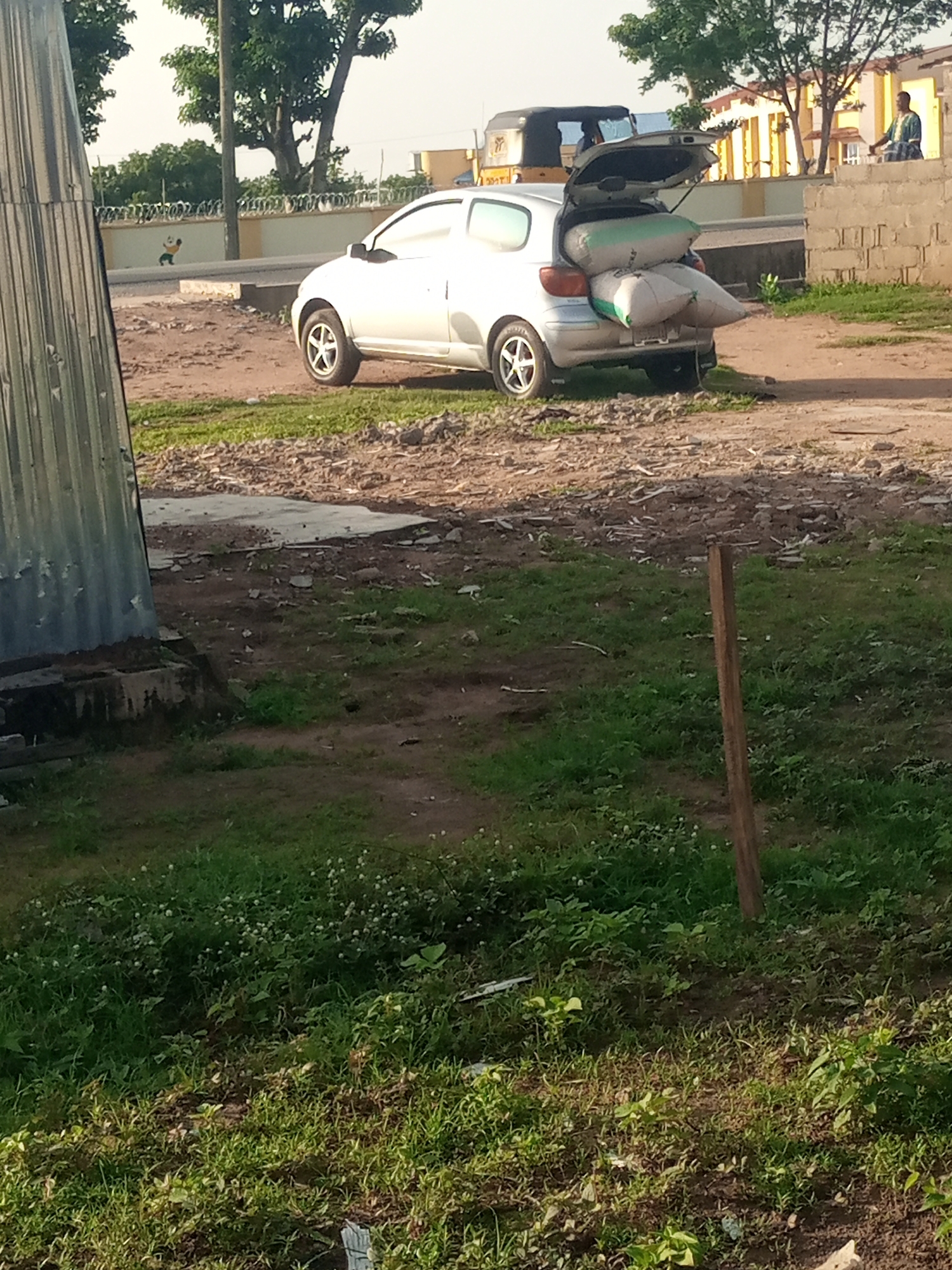 A white hatchback car is parked on a dirt road in Jalingo, Nigeria. The trunk is open and filled with several large sacks. A man in a blue and white garment stands to the right of the car, near a brick wall. In the background, there is a yellow building and a three-wheeled vehicle. The scene is an outdoor environment with sparse vegetation and debris on the ground.