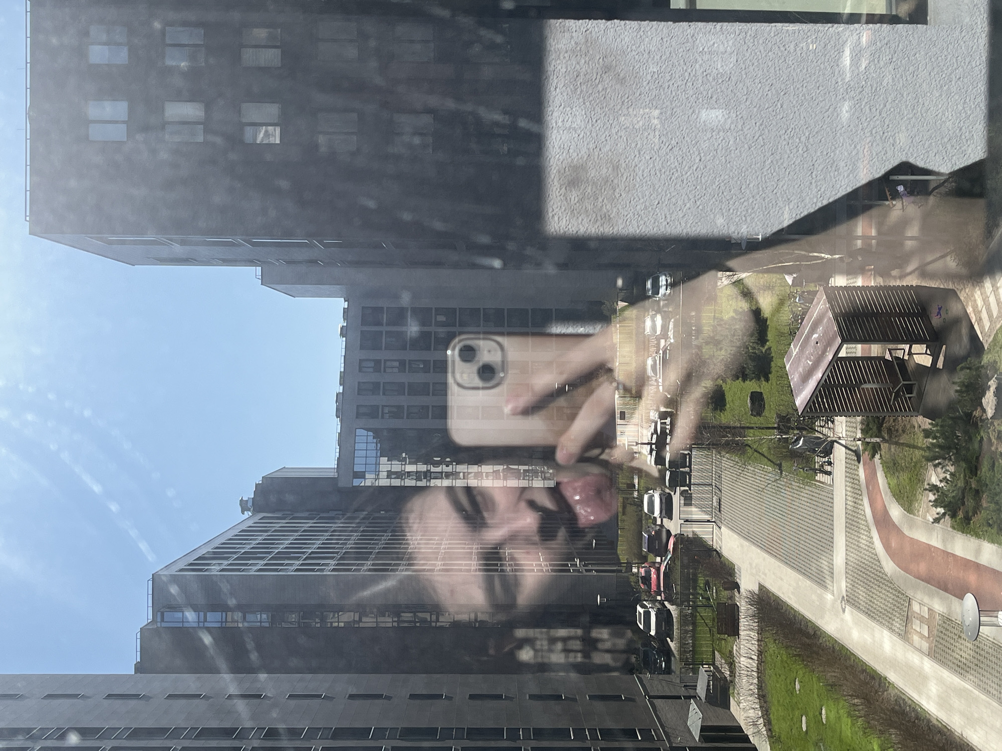 The image depicts a reflection in a window, showing a view of an urban environment. Tall, modern apartment buildings with rows of windows dominate the cityscape, suggesting a residential area. A street below features parked cars, pedestrian walkways, and landscaped areas with grass and trees. Reflected in the window is a person holding a smartphone, taking a picture. Their face and outstretched tongue are visible, superimposed over the cityscape. The overall scene appears to be taken on a sunny day in a modern urban setting. Based on the style of architecture, street design and parked vehicles visible in the image, it is possibly taken in Saint Petersburg, Russia.