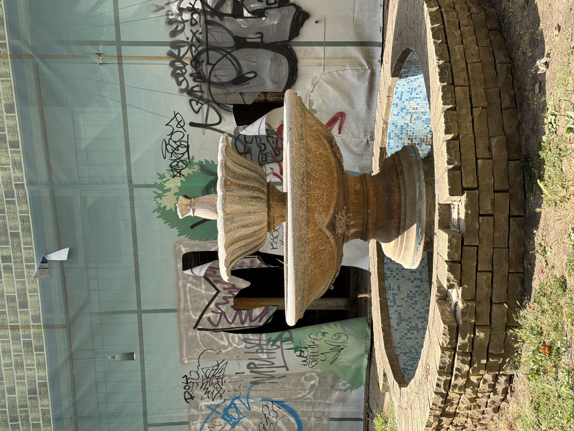 The image shows a tiered, stone fountain with a blue tiled basin, situated in what appears to be an urban outdoor area. The fountain is surrounded by a low, stone wall and dry grass. Behind the fountain, a large, light blue tarp is stretched across a metal frame, obscuring the background. The tarp is covered with graffiti in various colors, including black, white, blue, and red. A single, bare lightbulb hangs from a wire in front of the tarp. The overall impression is one of neglect or ongoing construction, with the graffiti suggesting an unauthorized artistic presence. The location is identified as Rostov-on-Don, Russia, but no specific landmarks or context are provided to further pinpoint the exact site. There are no people or animals visible in the image.