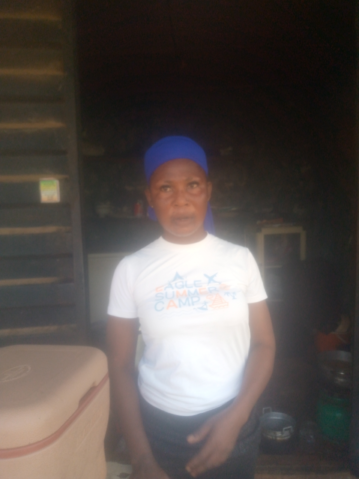 A woman with a blue head covering stands in a doorway. She is wearing a white t-shirt with the words "Eagle Summer Camp" on it. To her left is a container, and behind her in the dark interior are various items including a table, pots, and containers. The location is Unknown, Unknown.