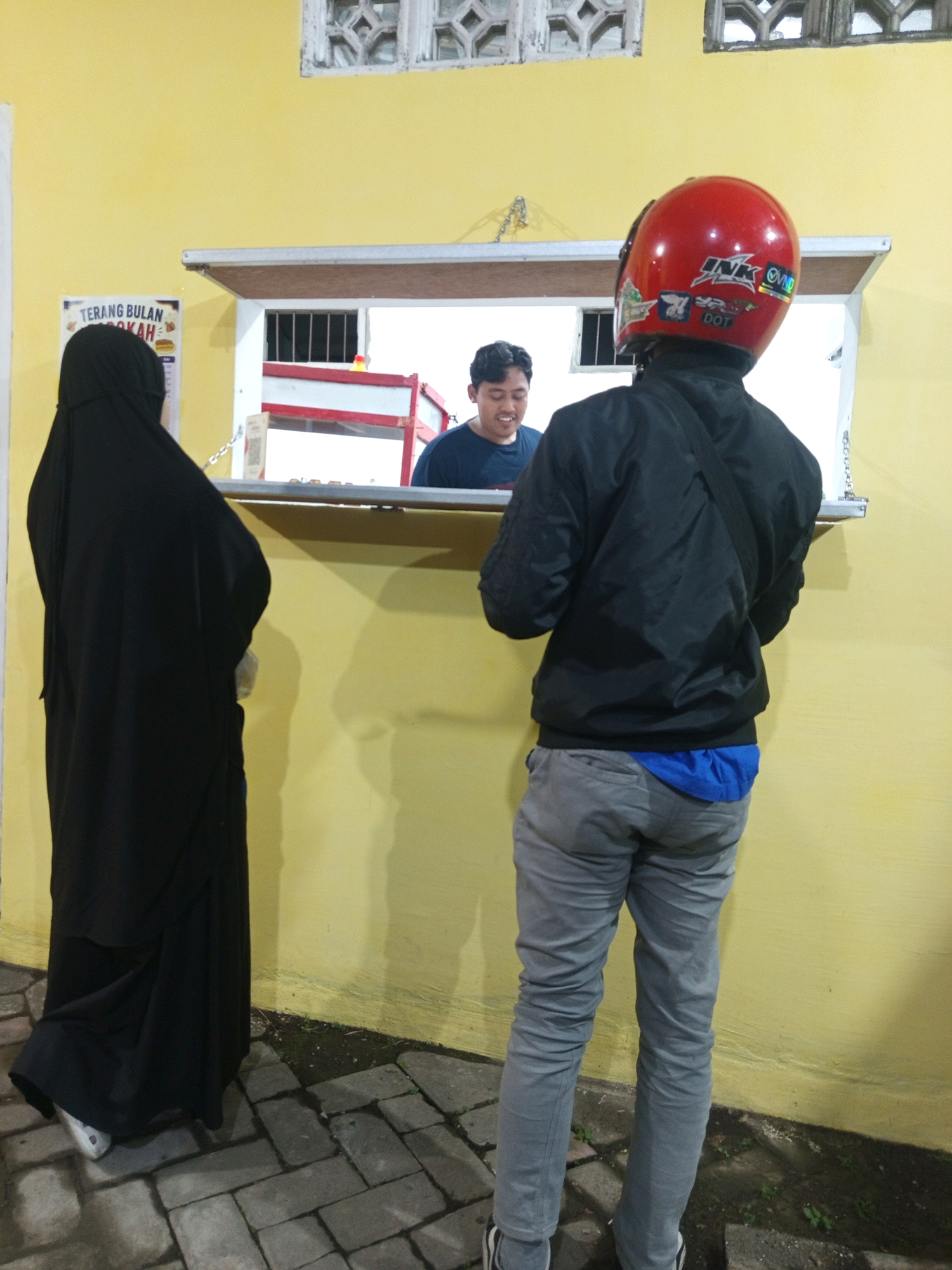 In Singosari, Indonesia, a person wearing a black abaya and hijab stands facing a yellow wall with a serving window. Behind the window, a person in a blue t-shirt is visible. In front of the window, a person wearing a red helmet, black jacket, and grey pants faces away from the viewer, towards the serving window. The scene appears to be an outdoor food stall or small eatery. The ground is paved with grey interlocking bricks, and some grass is visible along the base of the wall. The serving window has a white frame and a metal grate on the inside. A white sign with text, partially obscured by the person in the abaya, is mounted on the wall to the left of the serving window. A chain hangs from the top of the wall to the left of the serving window.