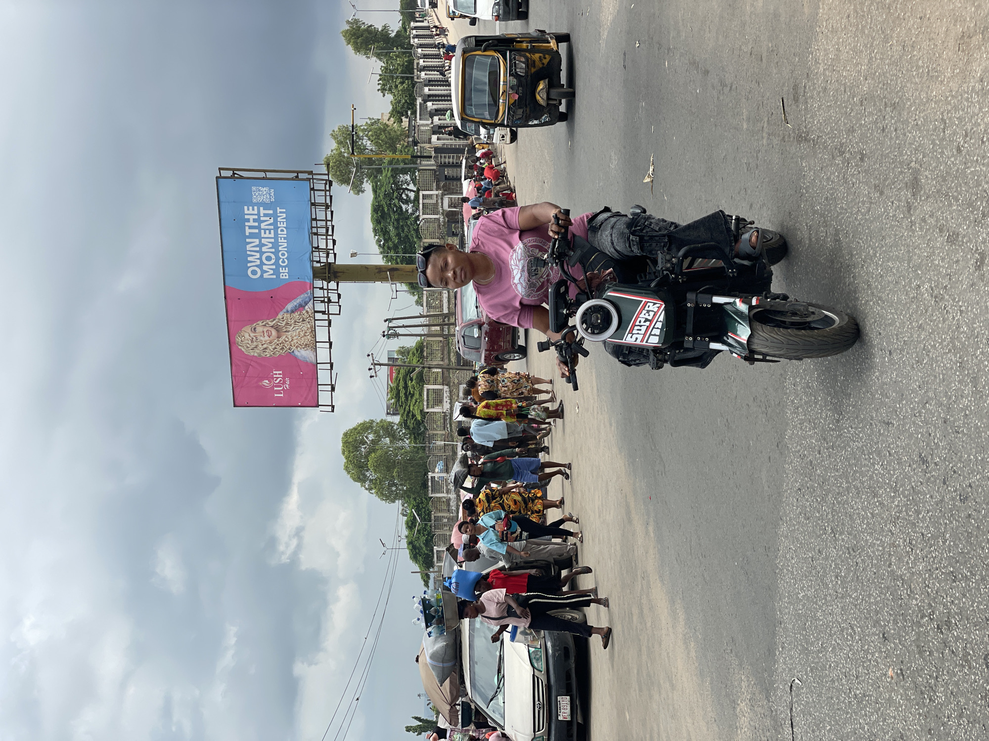 This image depicts a street scene in Onitsha, Nigeria, on a cloudy day. In the foreground, a woman is riding a green and black electric motorcycle. She is wearing a pink t-shirt with a graphic design and dark, textured pants. The motorcycle has the word "SUPER" emblazoned on its front in white letters with a red outline.
In the middle ground, a bustling street is visible with a variety of people and vehicles. A white car is parked on the left, loaded with items on its roof, including bottles of water. Several people are walking on the street, some carrying items on their heads. To the right, a yellow auto-rickshaw is driving down the road. Further in the background, there is a large billboard advertising "LUSH Hair" with the slogan "OWN THE MOMENT BE CONFIDENT" and a QR code. Trees and buildings are visible behind the billboard, and a white patterned fence runs along the right side of the road. The overall environment suggests a busy urban commercial or transit area.