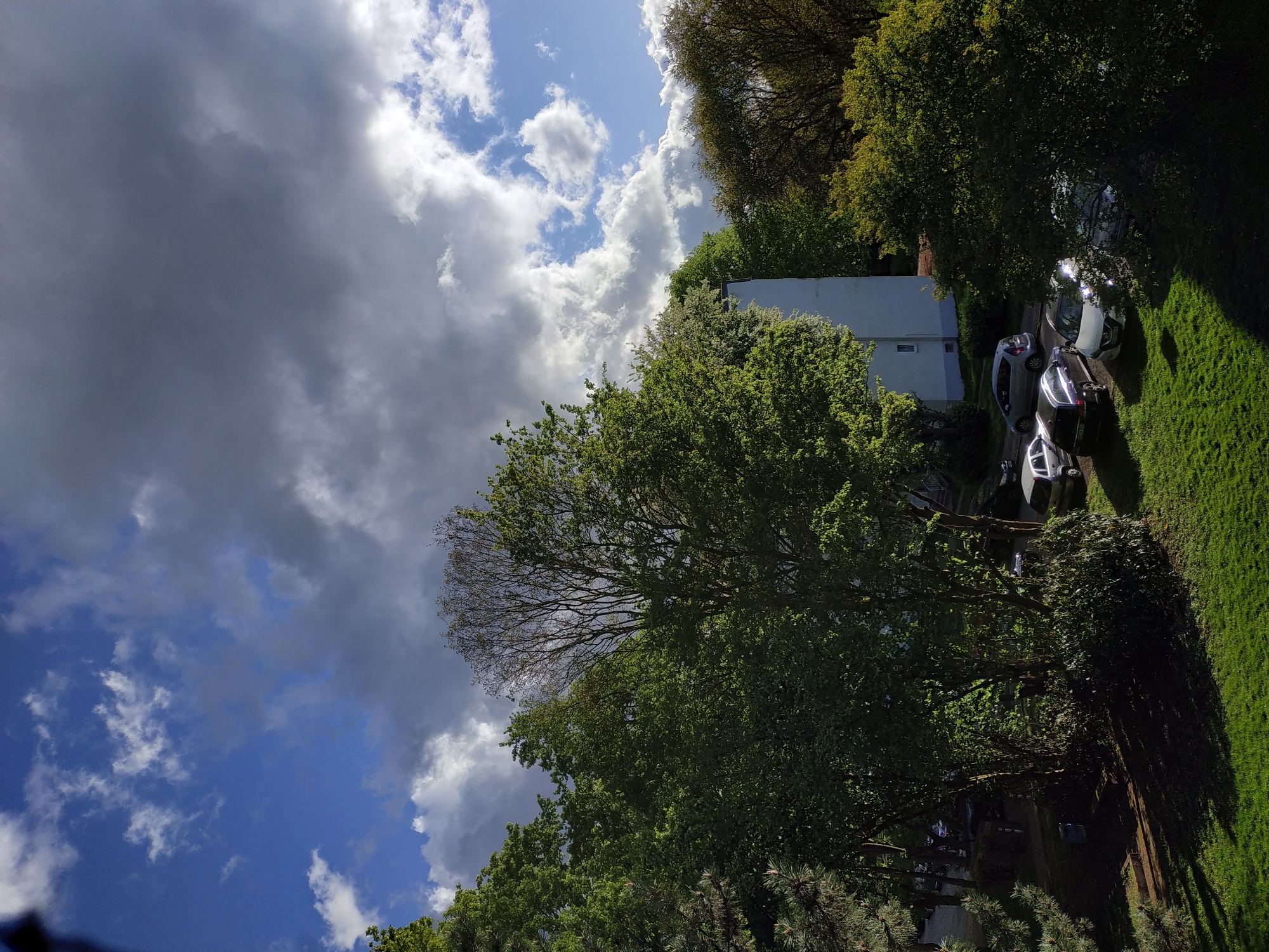 The image shows an outdoor scene, likely residential, in daylight. The sky is partly cloudy, with a mix of dark storm clouds and patches of blue sky with white cumulus clouds. In the foreground and midground, there are various trees with green foliage. The background reveals a light-colored building and several parked cars on what appears to be a driveway or roadside. There is also a strip of green grass. Without further information, it's impossible to confirm this location is in Dortmund, Germany.