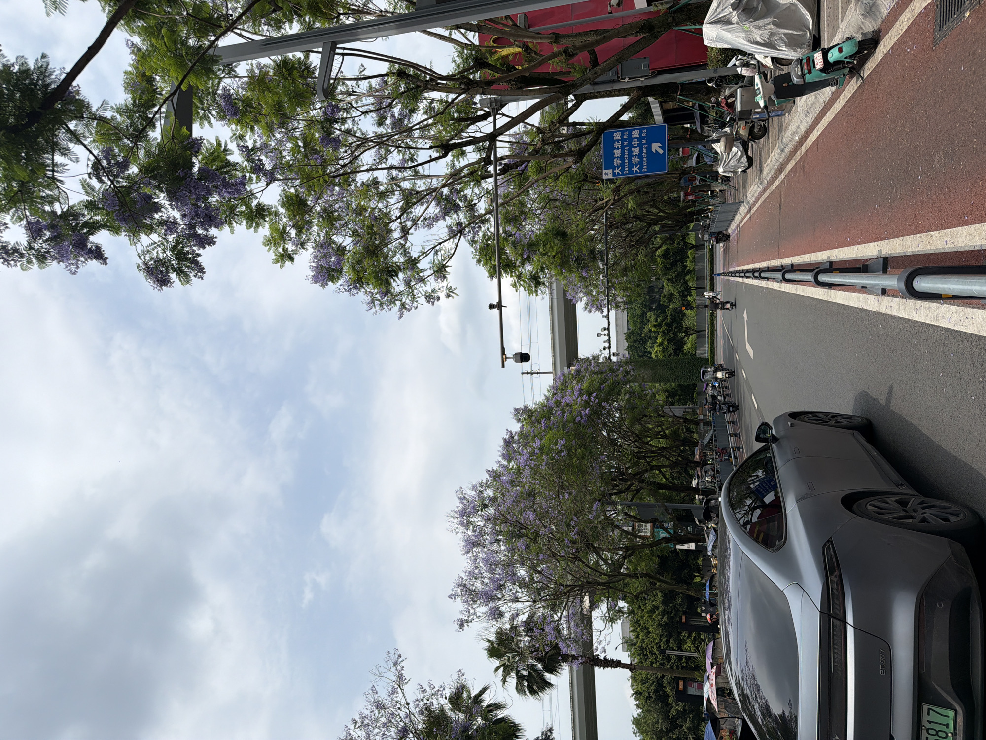 The image depicts a street scene in Chongqing High-tech Zone, China. The view is taken from a roadside, showing a multi-lane road with a red-colored pedestrian/bicycle path on the right. Jacaranda trees with purple blossoms line the street, partially obscuring the sky which is lightly clouded. A light grey car is parked on the right side of the road in the foreground. Further down the road, parked electric scooters are visible. A blue street sign displaying road names in Chinese characters, including "Daxuecheng N. Rd." and "Daxuecheng M. Rd.", is fixed to a pole along the road. Traffic lights can be seen hanging overhead. The car has a license plate with Chinese characters and the identification "et007." In the background, there's an elevated road structure.