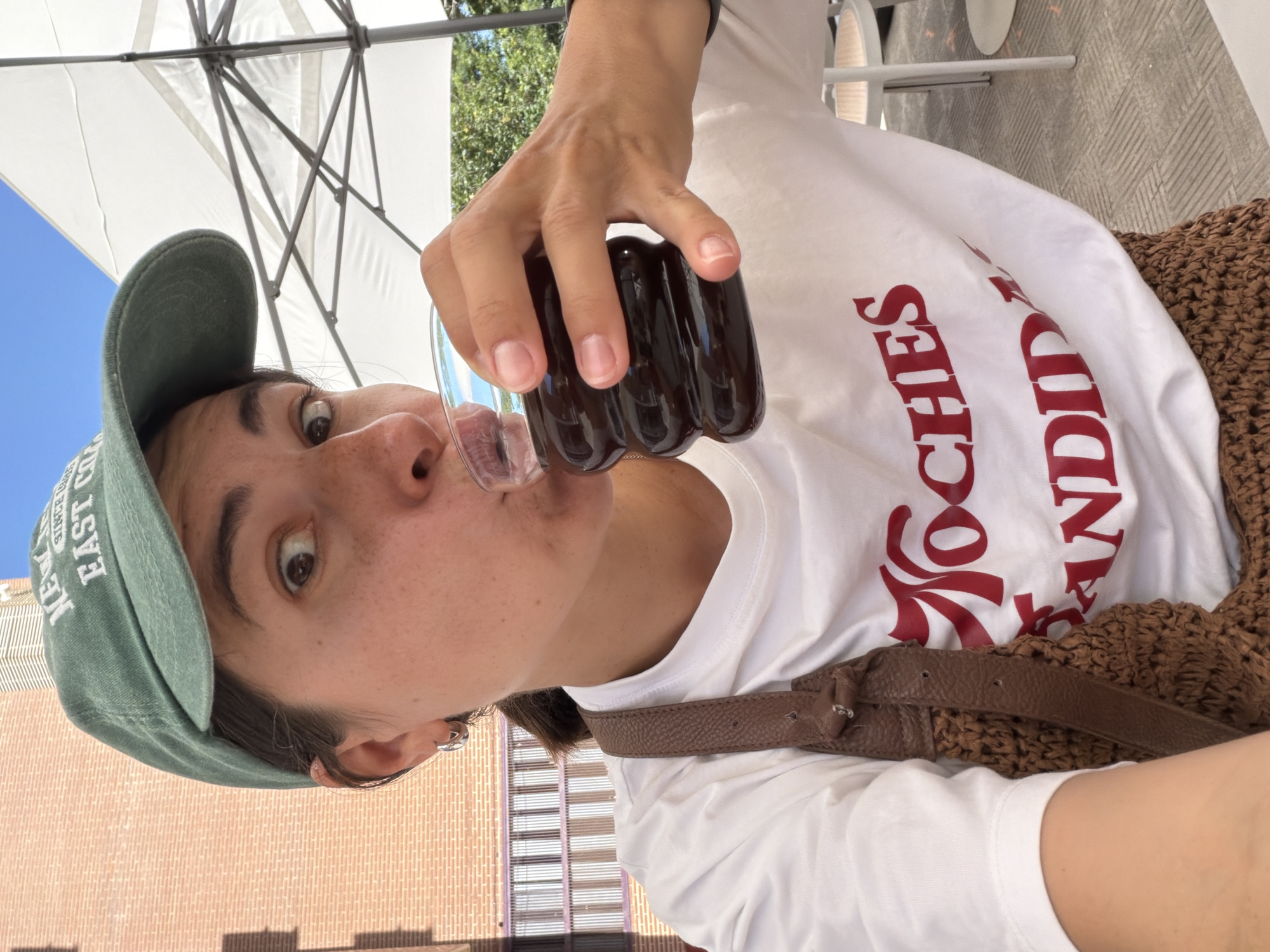 A medium shot captures a woman wearing a green baseball cap and a white t-shirt with red text reading "Noches Candidas." She is holding a ribbed glass of dark liquid and drinking from it. The woman has brown hair and is wearing silver hoop earrings. She has a brown woven bag with a leather strap slung over her shoulder. In the background, a brick building and a white umbrella are visible. The scene appears to be outdoors, possibly at a cafe or restaurant, with tables and chairs in the periphery. The lighting suggests it is daytime. The background building's architecture and the presence of a white umbrella are consistent with outdoor dining establishments found in cities like Valencia, Spain.