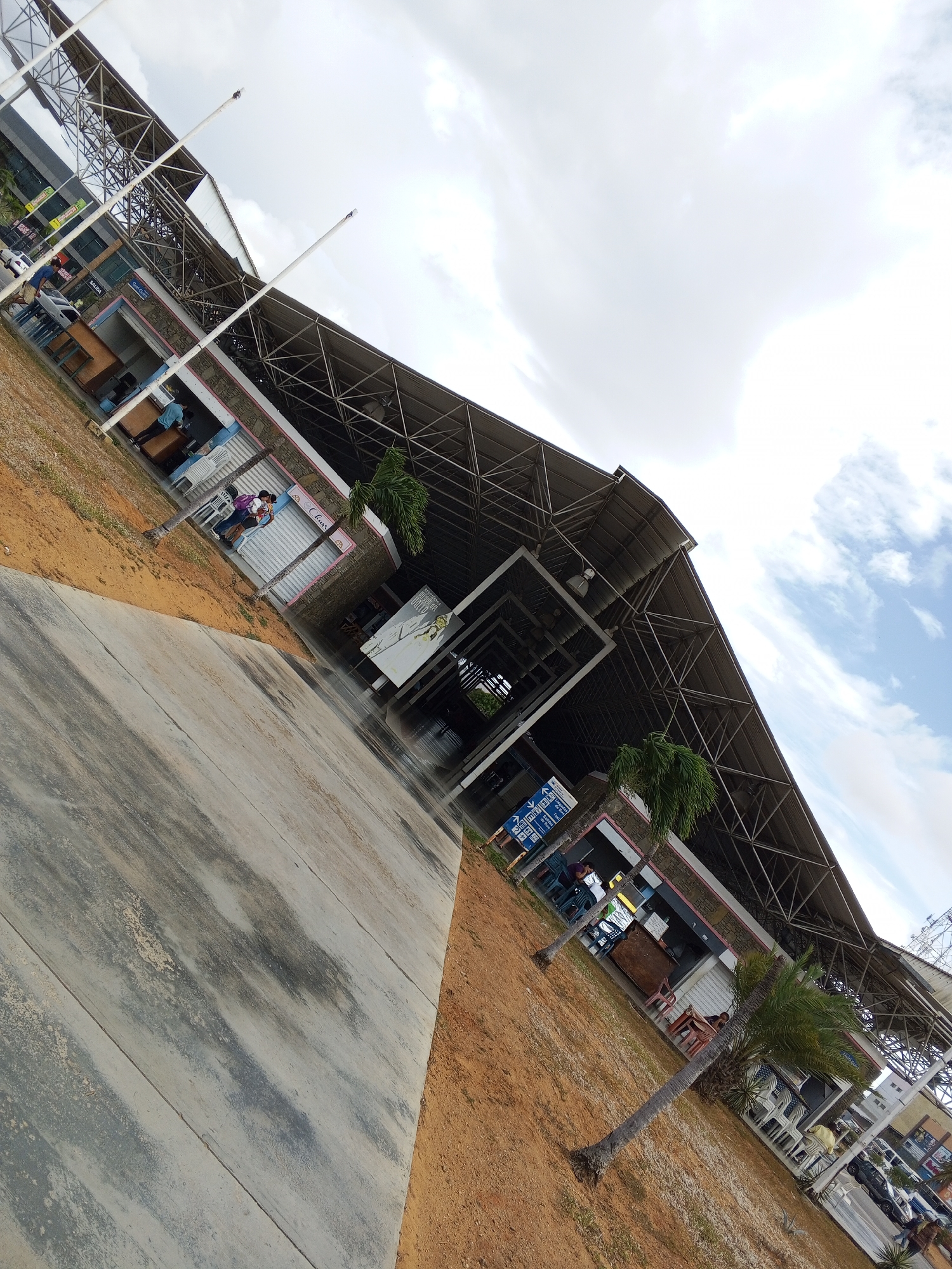 Captured from a slanted perspective, the image showcases a large structure, possibly a market or transportation hub, located in an urban setting. The building features an extensive metal framework supporting a broad roof that spans over a shaded area. Along the sides, numerous small kiosks or shops line the perimeter, some of which are occupied by people, hinting at commercial activity. The ground around the structure is a mix of paved surfaces and reddish-brown soil, with a few palm trees adding a touch of greenery. In the distance, other buildings and vehicles are visible, indicating a populated area. The sky above is partly cloudy, suggesting either early morning or late afternoon lighting conditions.