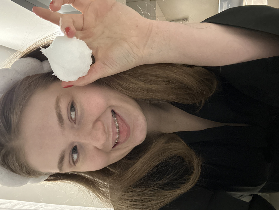 A fair-skinned young woman with light brown hair smiles at the camera, her teeth visible with braces. She holds a ball of white cotton above her head with her right hand, nails painted red. She wears a white fluffy headband with cloud shapes. A black garment with a low neckline is also visible. The photo is taken indoors and the image is rotated sideways.