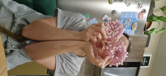 An adult individual is captured indoors, likely within a domestic kitchen setting in Kudrovo, Russia. The subject's face is partially obscured by two pink-frosted donuts, heavily garnished with small white marshmallows, held directly in front of her face with both hands. Her lips are visible between the donuts, pursed as if in a kissing gesture. Her forehead and the top of her head are visible above the donuts. She wears a grey short-sleeved t-shirt, and a gold-colored ring is present on the ring finger of her left hand. Her arms are bent, and her forearms are visible. Long, dark hair extends from behind her t-shirt on the lower right. The background features a stainless steel refrigerator door adorned with various magnets, including a blue rectangular one depicting two cartoon figures. Above the refrigerator, a green potted plant is visible. To the right, a wooden cabinet and a white object with partial Russian text ("ПРЕМЬЕР") are present. A kitchen appliance, possibly a microwave, is located above and to the right of the subject's head. A light-colored wooden surface, likely a table or countertop, is in the foreground, and a dark green object is partially visible on the lower left background.