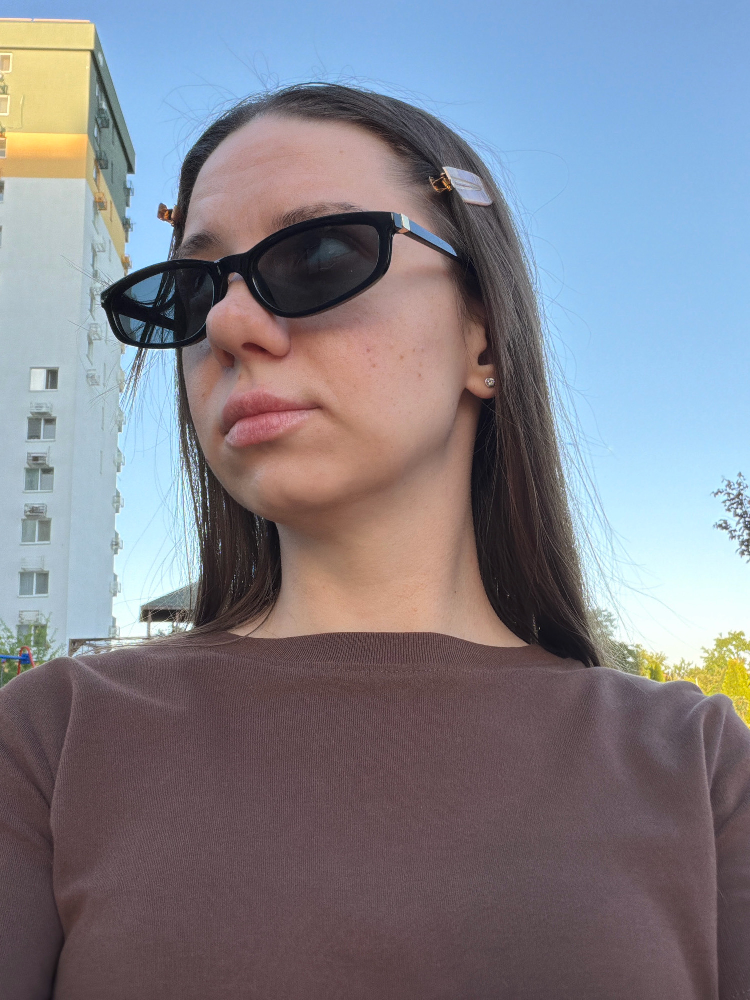 A young woman with long, dark brown hair, wearing black sunglasses and a brown long-sleeved shirt, is shown in a close-up, chest-up shot. She has a single stud earring in her visible ear. Two clear hair clips are visible in her hair, one on each side of her head. The background suggests an outdoor urban environment, with a tall, multi-story building, likely an apartment complex, to the left. The building is light-colored with visible window frames and air conditioning units. The sky is clear and blue. The overall scene is consistent with a sunny day in Kyiv, Ukraine, possibly in a residential area with buildings and some greenery.