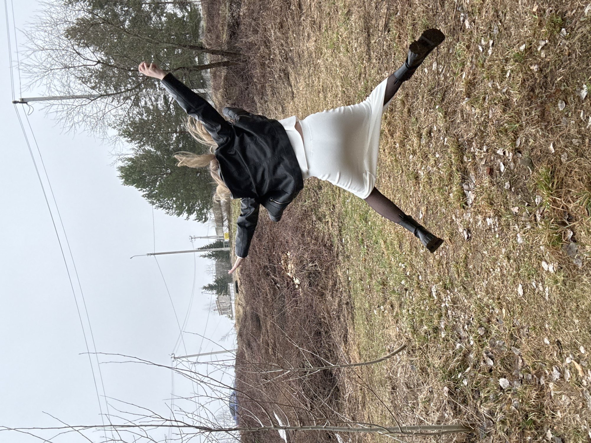A woman in a black leather jacket, white dress, and black tights is captured mid-air in what appears to be a celebratory or energetic pose, with arms outstretched and legs spread. She is wearing black boots. The location is a grassy area interspersed with fallen leaves and dry brush, suggesting a transitional season like late fall or early spring. There are bare trees and shrubs in the background. In the distance, there are buildings, power lines, and evergreen trees, indicating an urban or suburban setting. The image seems to have been taken on an overcast day. The scene is potentially located in Chaikovsky, Russia.