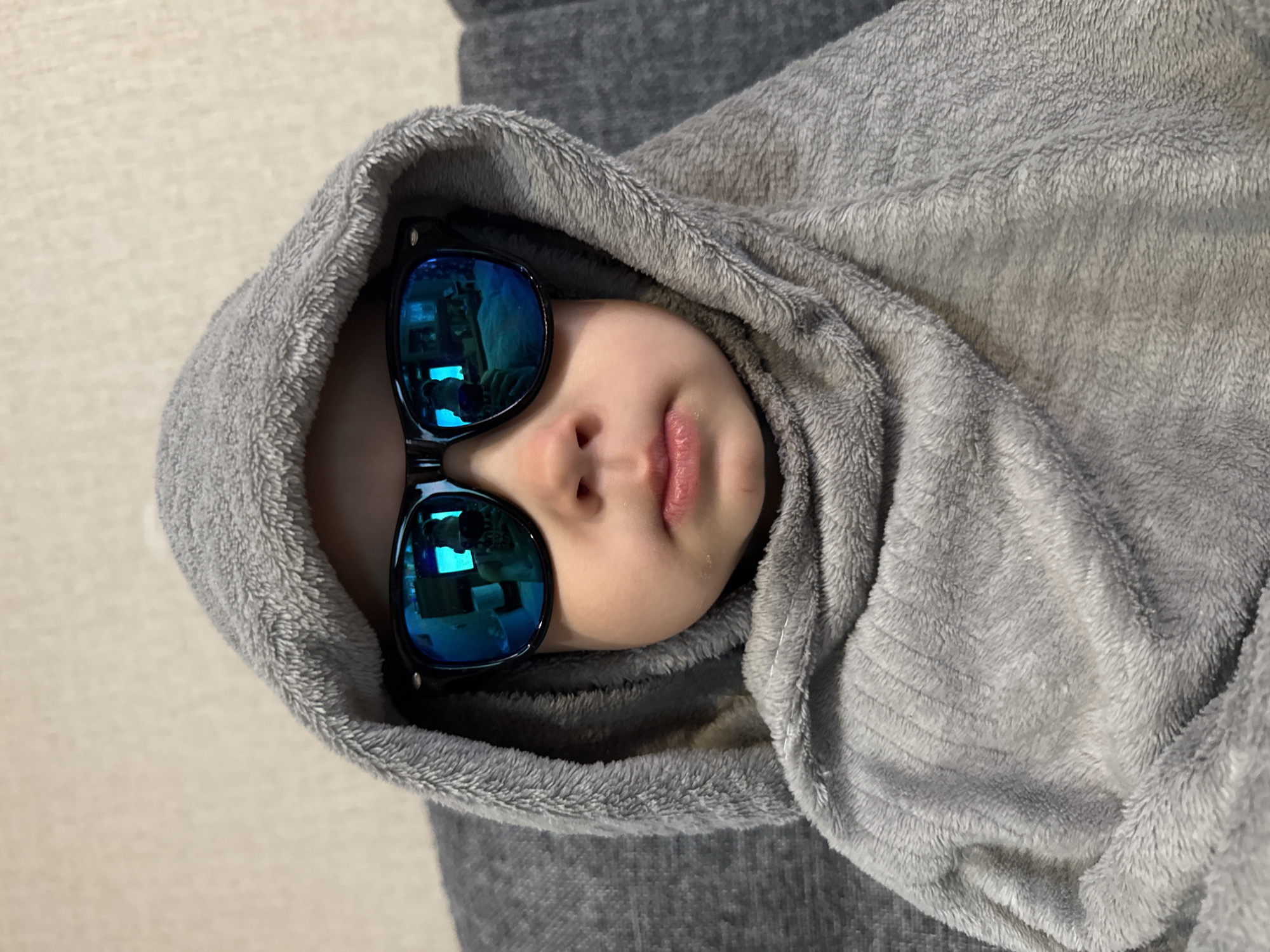 A child lies swathed in a gray, hooded blanket, face towards the camera. The child is wearing large, black sunglasses with reflective blue lenses. The background is a light-colored floor and gray fabric. There is no indication of Rusanovo, Russia.