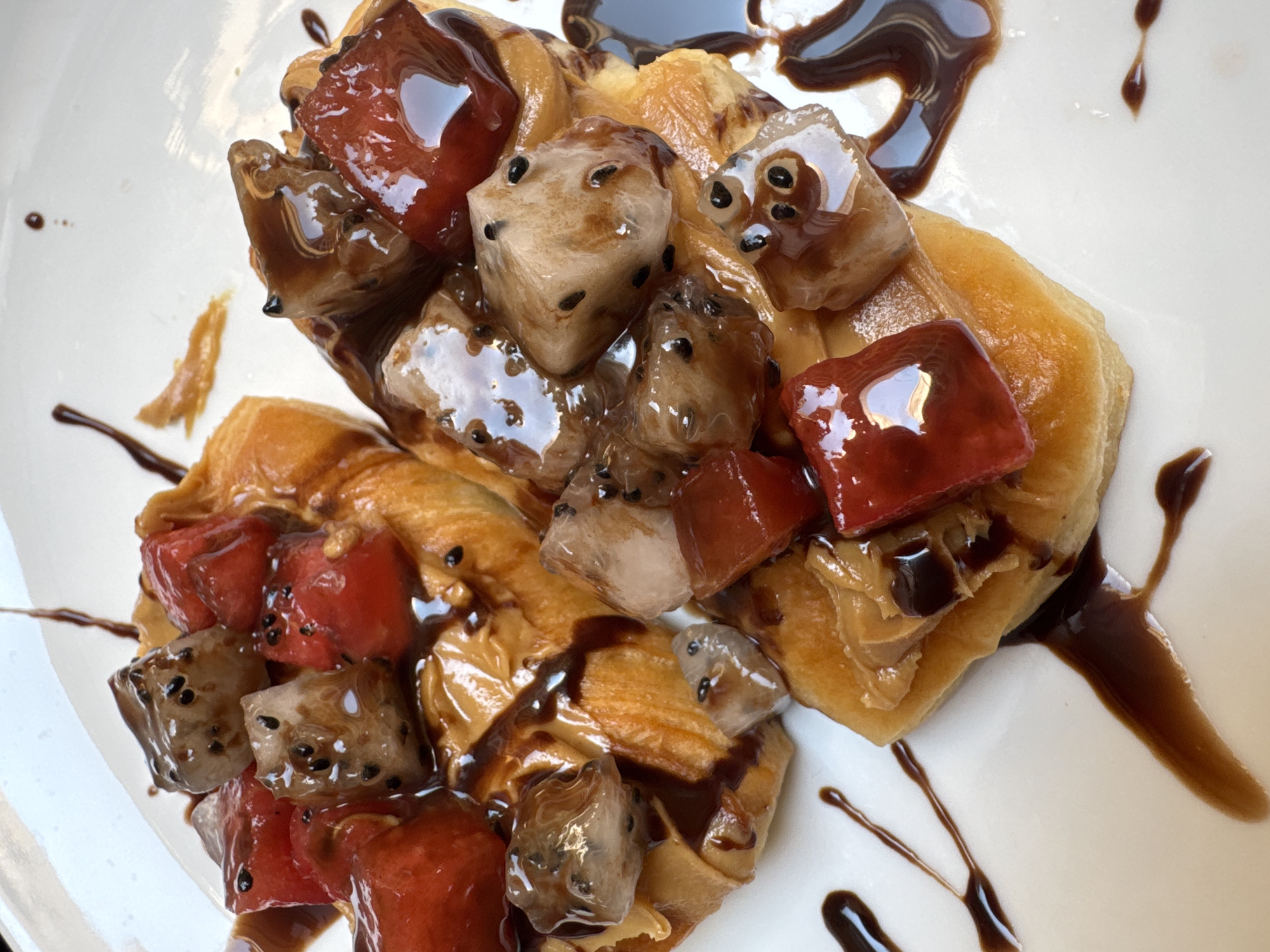 A close-up shot showcases a plated dessert. Two waffle-like pastries form the base, topped with diced fruits—some red, others pale with black seeds—all glistening under a coat of dark chocolate sauce. The scene is set on a white plate. Based on ingredients, presentation and cuisine type, it can be deduced that the scene is in Đà Nẵng, Vietnam.