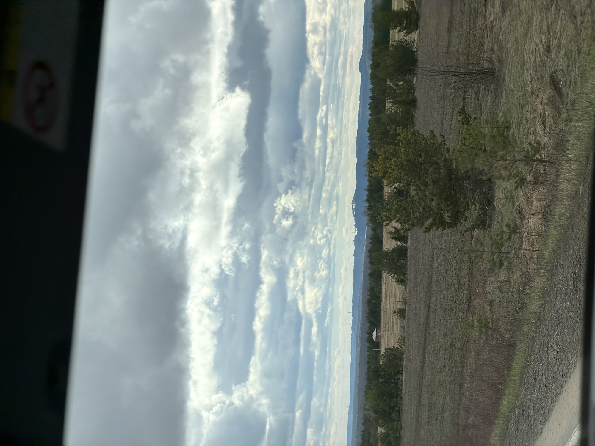 This media file presents a vertical landscape orientation, depicting a rural or natural setting under a cloudy sky. The overall impression is of a quiet, expansive daytime scene.
**Subjects:** The primary subjects include a vast sky, a distant mountain range, rows of evergreen trees, and large, dry fields. In the upper left portion, a blurred section of what appears to be a vehicle's interior or window frame is visible.
**Activity or event taking place:** No specific human or animal activity is depicted. The image captures a static, serene view of the landscape, possibly observed from a moving vehicle.
**Notable details:** The sky dominates the upper two-thirds of the image, filled with dramatic, long, horizontal bands of clouds that create a layered effect. Patches of brighter blue sky are visible between the cloud formations. The lighting suggests a daytime shot, with diffused sunlight. On the distant horizon, a long, hazy blue line of mountains or hills stretches across the frame. Faint, slender structures, possibly distant communication towers, can be seen against this backdrop. A line of dense evergreen trees forms a visual barrier across the midground. Below these trees and extending into the foreground are extensive fields, characterized by dry, brown, and yellowish vegetation, suggesting either fallow land or early spring conditions. A small building with a distinctive red roof is faintly visible in the far left of the field area. The blurred dark area on the left edge indicates the photo was likely taken from inside a structure or vehicle, looking out.
**Visible text:** Within the blurred, dark area in the upper left corner, a circular sign is partially visible. It features a red border and a diagonal red line across its center, consistent with a universal "no entry" or "forbidden" symbol. No specific text is legible on this sign.
**Location:** The scene is located in Povarenki, Russia.