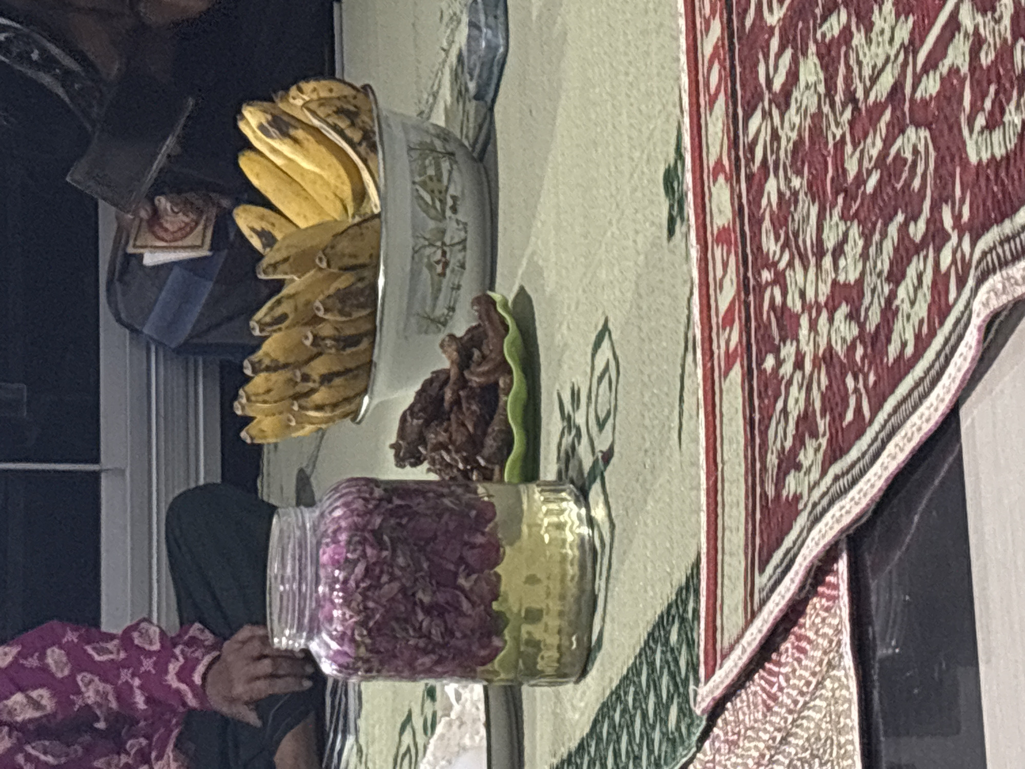 The image depicts a dimly lit indoor scene, likely a home in Tirtomoyo, Indonesia. In the foreground, a woven mat with a red and cream geometric pattern is visible. To the left, a person is seated cross-legged, wearing a purple patterned garment. In front of the seated person is a clear glass jar filled with what appears to be dried rose petals and a golden liquid, possibly rosewater or a similar infusion. To the right of the jar, a green plate holds a pile of dates. Further to the right, a white bowl contains a bunch of bananas. The background shows a dark surface that could be a table or floor, and hints of furniture or a window. The overall impression is one of a domestic setting, possibly for a gathering or meal.