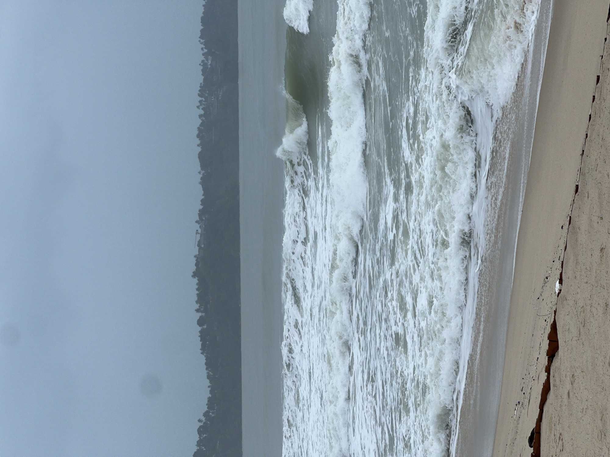 The image shows a beach scene. Waves are crashing onto the shore, with white foam visible. The beach is sandy, and there's a landmass with trees in the background. The sky is overcast, suggesting a cloudy or foggy day. There are no people visible in the scene. The location is identified only as Unknown, Unknown.