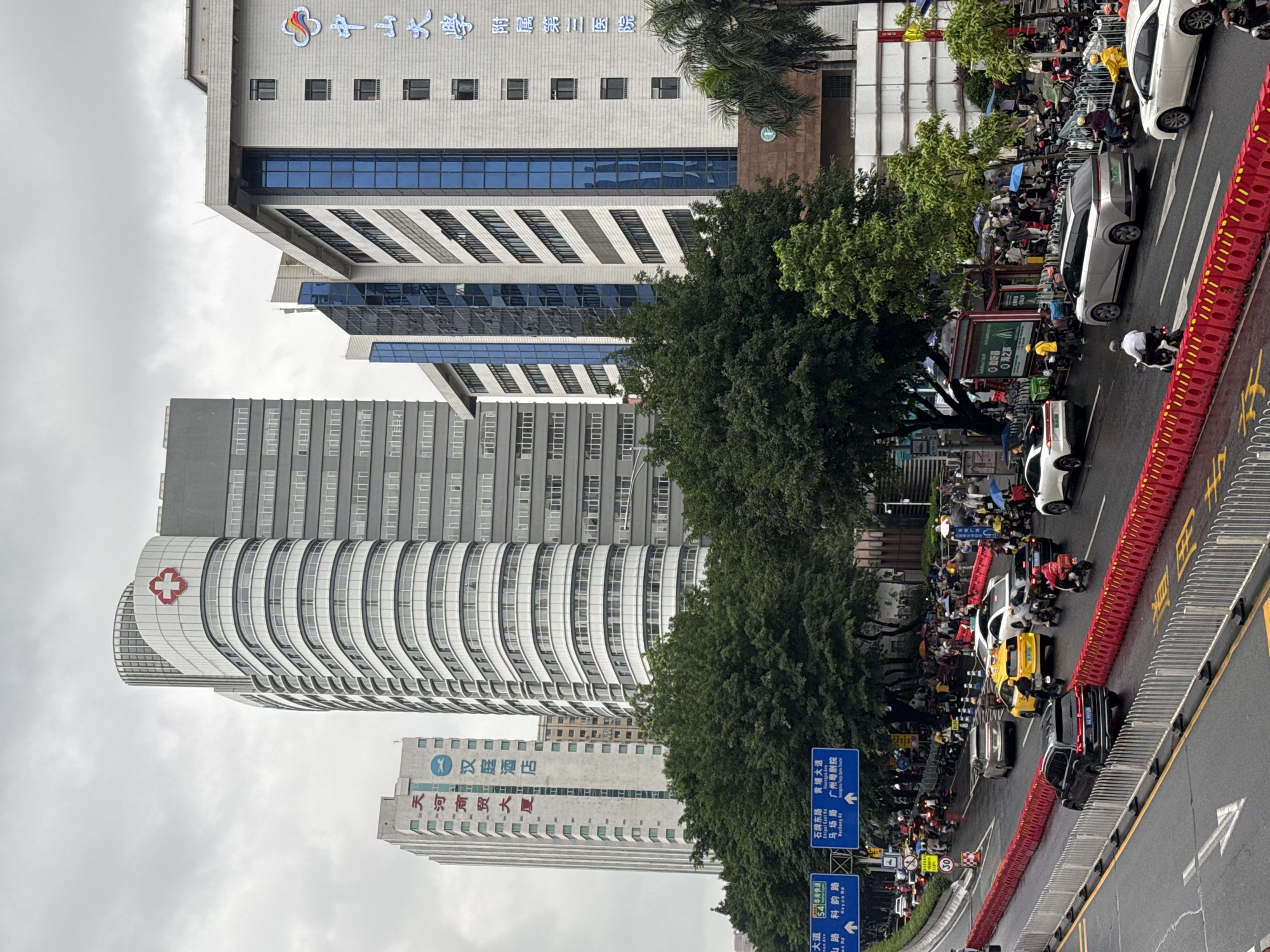 The image captures a scene in Tianhe District, China. Tall buildings, including a curved building with a red cross, rise against a cloudy sky. Street-level activity includes a line of cars and people walking alongside the road, some carrying umbrellas. Road signs are visible, directing traffic to various locations, including “Shipai East Rd”, and “Keyun Rd”. A red and white barrier runs along the roadside. The scene appears to be an urban street with a blend of architectural structures and pedestrian traffic.