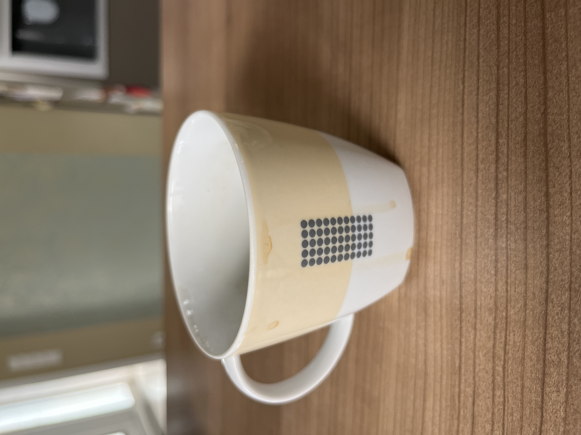 A coffee mug with a tan band and a pattern of dark grey dots sits on a wooden table. The mug has some brown stains on its exterior, suggesting it has been used. The background is blurred, showing a window on the left and a kitchen appliance on the right. The scene appears to be indoors, possibly in a home or a cafe. The image does not provide any information about the location in the city of Bad Laer, Germany.