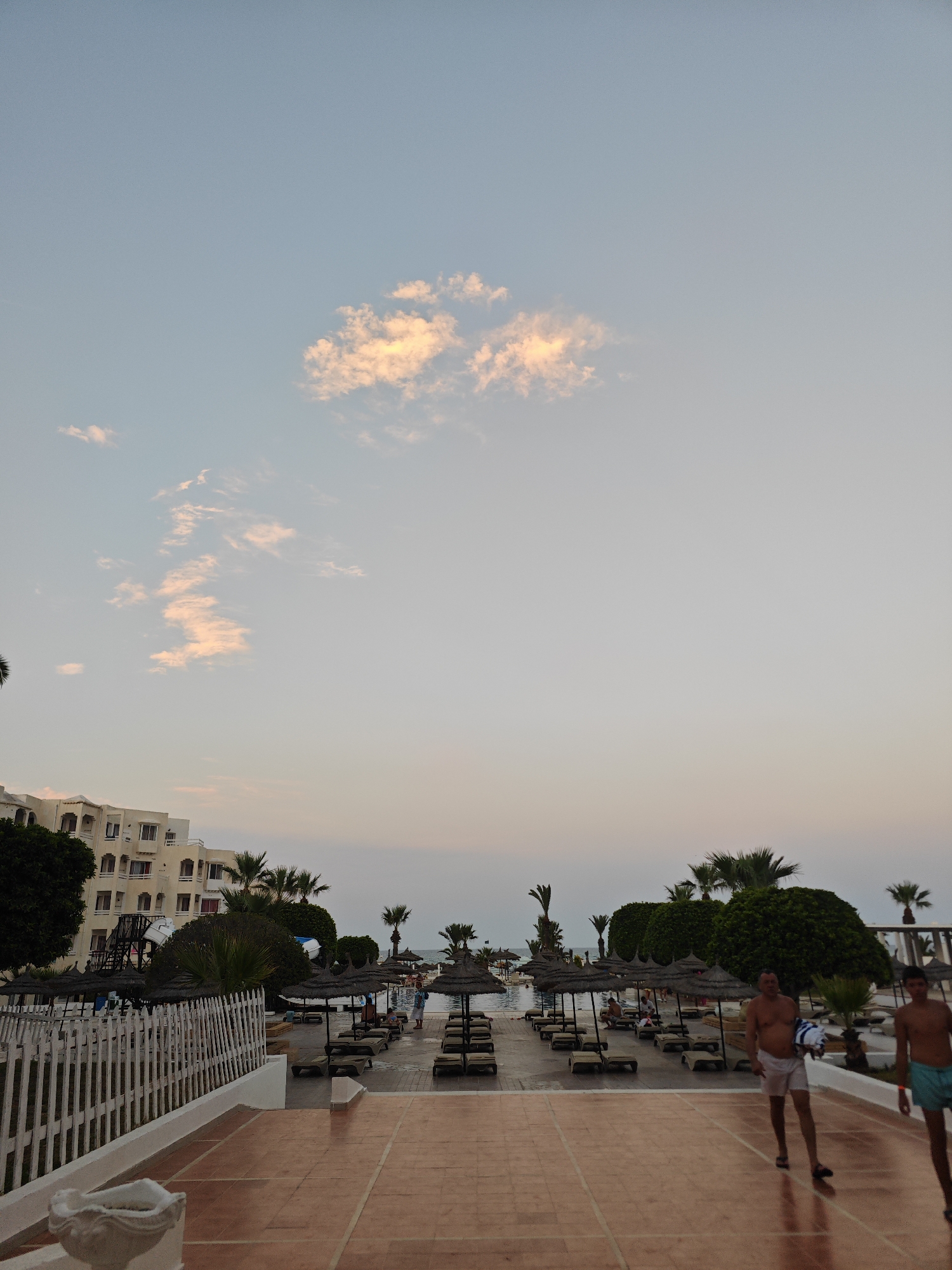 The image depicts an outdoor resort scene, likely in Hiboun, Tunisia, during what appears to be late afternoon or early evening, judging by the soft lighting and pastel colors in the sky. In the foreground, a tiled pathway leads towards a white fence. Beyond the fence, rows of lounge chairs and thatched umbrellas are arranged on a sandy or paved area, suggesting a pool or beachside setting. A multi-story hotel building with balconies and windows is visible on the left side of the frame, nestled amongst trees and palm trees. Several people are present in the scene; two men are walking towards the foreground on the tiled path, one holding a towel. Other individuals can be seen near the pool or lounging under umbrellas in the middle ground. The sky above is a pale blue with soft, illuminated clouds. The overall atmosphere is tranquil and suggests a vacation or resort environment.