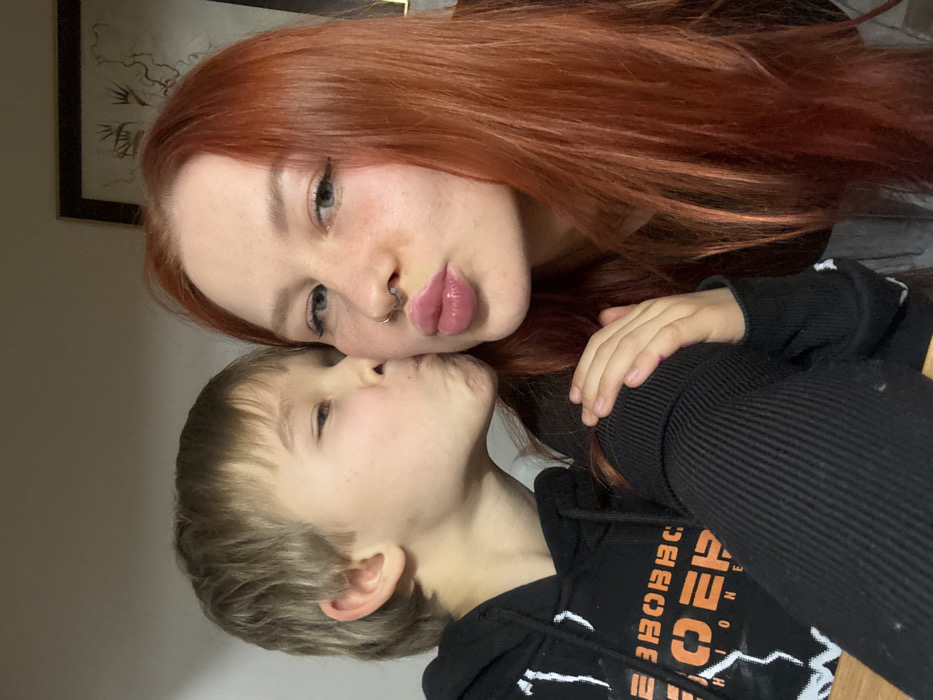 A woman with long, red hair and a nose ring puckers her lips toward a younger boy. The boy, with blonde hair, closes his eyes and leans in to kiss the woman on the cheek. The woman's right hand rests on the boy's shoulder, who is wearing a black hooded sweatshirt with white lightning bolt designs and orange lettering. A framed artwork is partially visible in the background, positioned above the woman's head.
