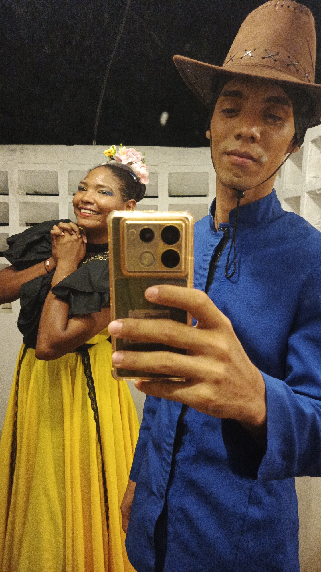 The image depicts two individuals in what appears to be Playa El Agua, Venezuela. A person wearing a brown cowboy hat and a blue shirt is holding a smartphone with a gold-colored case, presumably taking a picture or recording a video. Slightly behind and to the side of this person stands another individual, a woman with flowers in her hair, blue eyeshadow, a black ruffled top, and a yellow skirt with black lace trim. She is smiling and has her hands clasped together. The background consists of a white wall with a repeating square pattern. The overall lighting is somewhat dim, and the scene has a candid, informal feel.