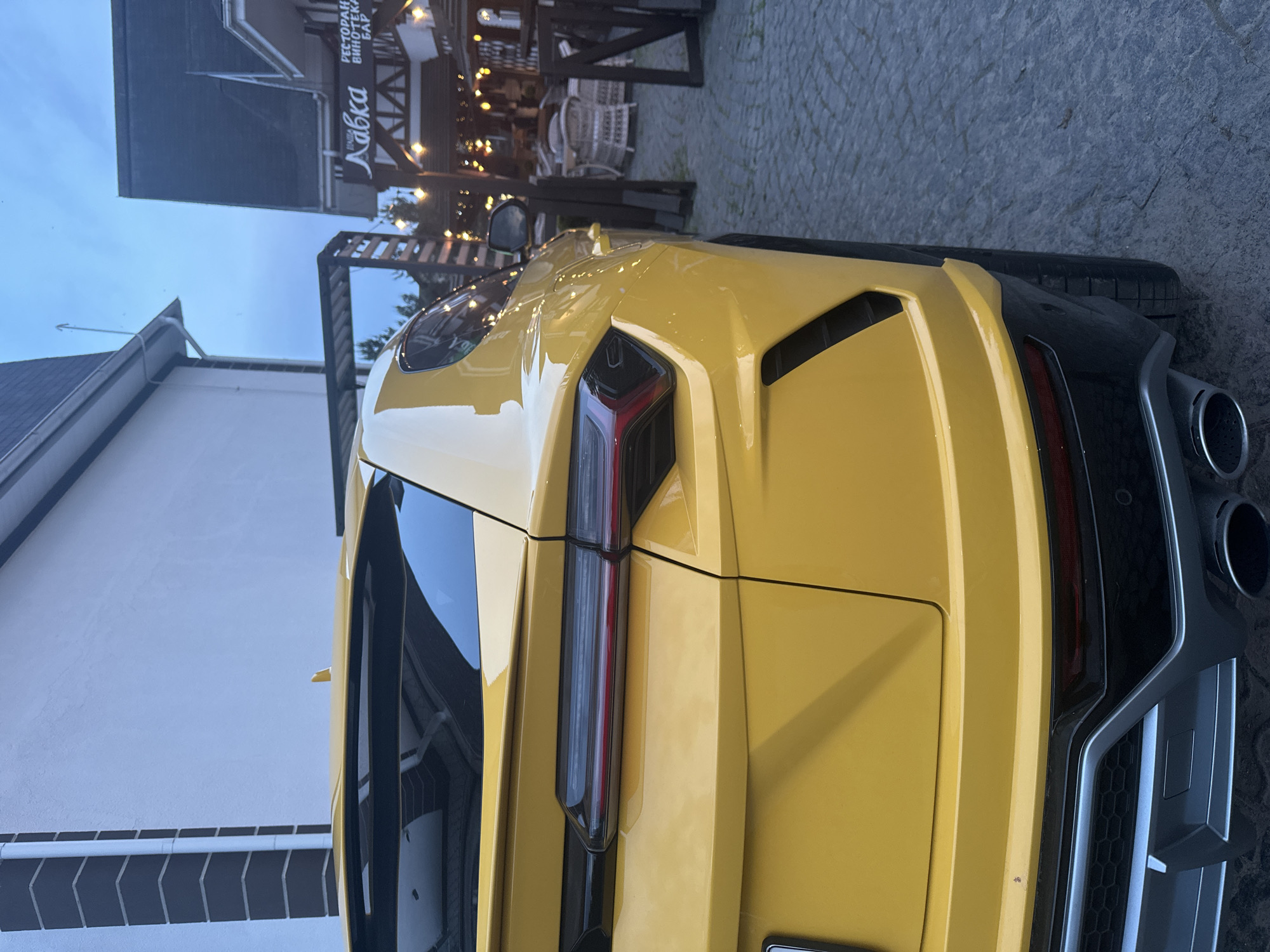 The image shows the rear of a yellow Lamborghini Urus parked on a paved surface, likely a street or parking area. The car's taillights and exhaust pipes are visible. In the background, there's a building with a sign written in Cyrillic script, appearing to read "Labka," and what seems to be the rest of the text reads "Pectopah Винотека Бар" which is most likely the name of a restaurant or bar. There is a building to the left with a white painted wall. The overall setting suggests a location in a city; based on the language of the sign, most likely Новоглаголево, Russia. The time appears to be dusk or early evening based on the sky.