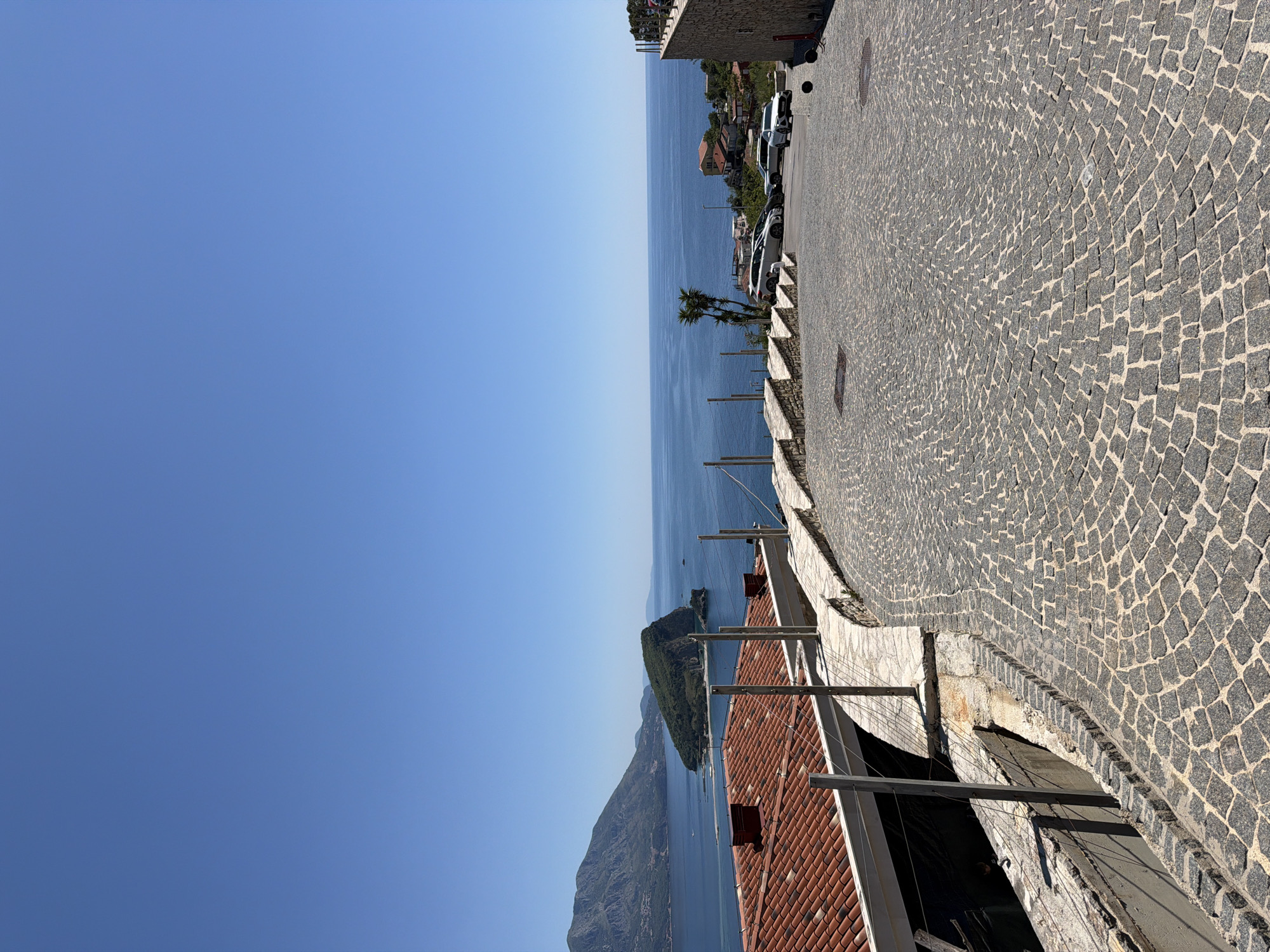 The image shows a view from a high point in or near Budva, Montenegro. The perspective looks out over the Adriatic Sea with clear blue skies above. An island covered in vegetation is visible in the middle distance. The foreground features a cobblestone street with a low wall and a rooftop with red tiles below it. The composition emphasizes the landscape and architectural elements. No people are visible in the frame. Several cars are parked in the background.