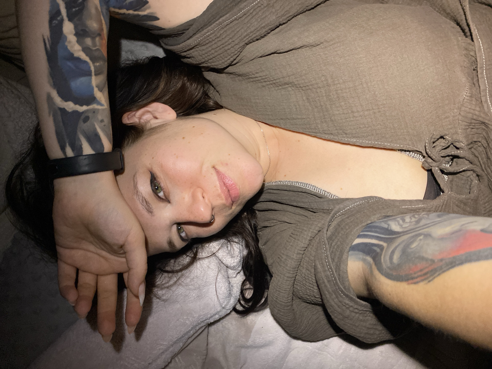 The image depicts a woman lying down with her head turned towards the camera. She is wearing a brown, textured top. Her right arm is extended across her forehead, partially obscuring her face. Tattoos are visible on both of her arms. Her eyes are open and looking directly at the camera. The background appears to be a bed or sofa with white bedding. The overall lighting is dim, suggesting an indoor setting. The image does not contain any specific information or visual cues that would allow for identification of the location as Chaikovsky, Russia.
