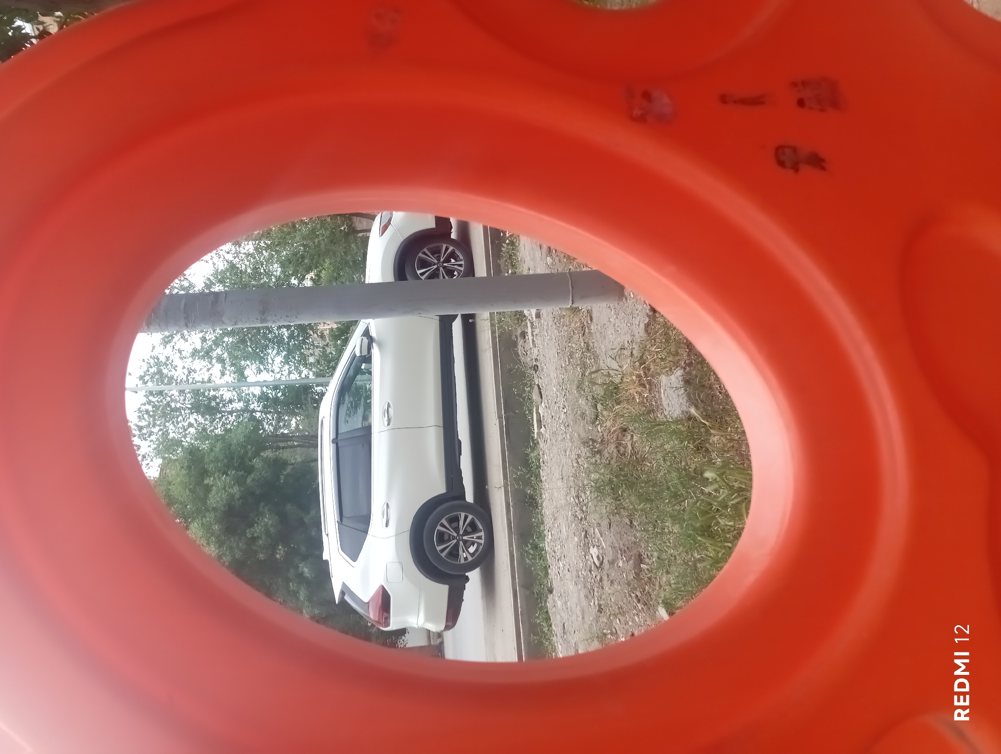 A white SUV is parked on the side of a road, partially obscured by an orange circular object in the foreground. The SUV is facing left, with its rear towards the viewer. The road appears to be paved, with a curb and some greenery alongside it. Trees and a utility pole are visible in the background. The orange object in the foreground has some faint sticker-like markings on it. The bottom left corner of the image displays "REDMI 12" in white text. The overall scene suggests an outdoor urban or suburban environment.