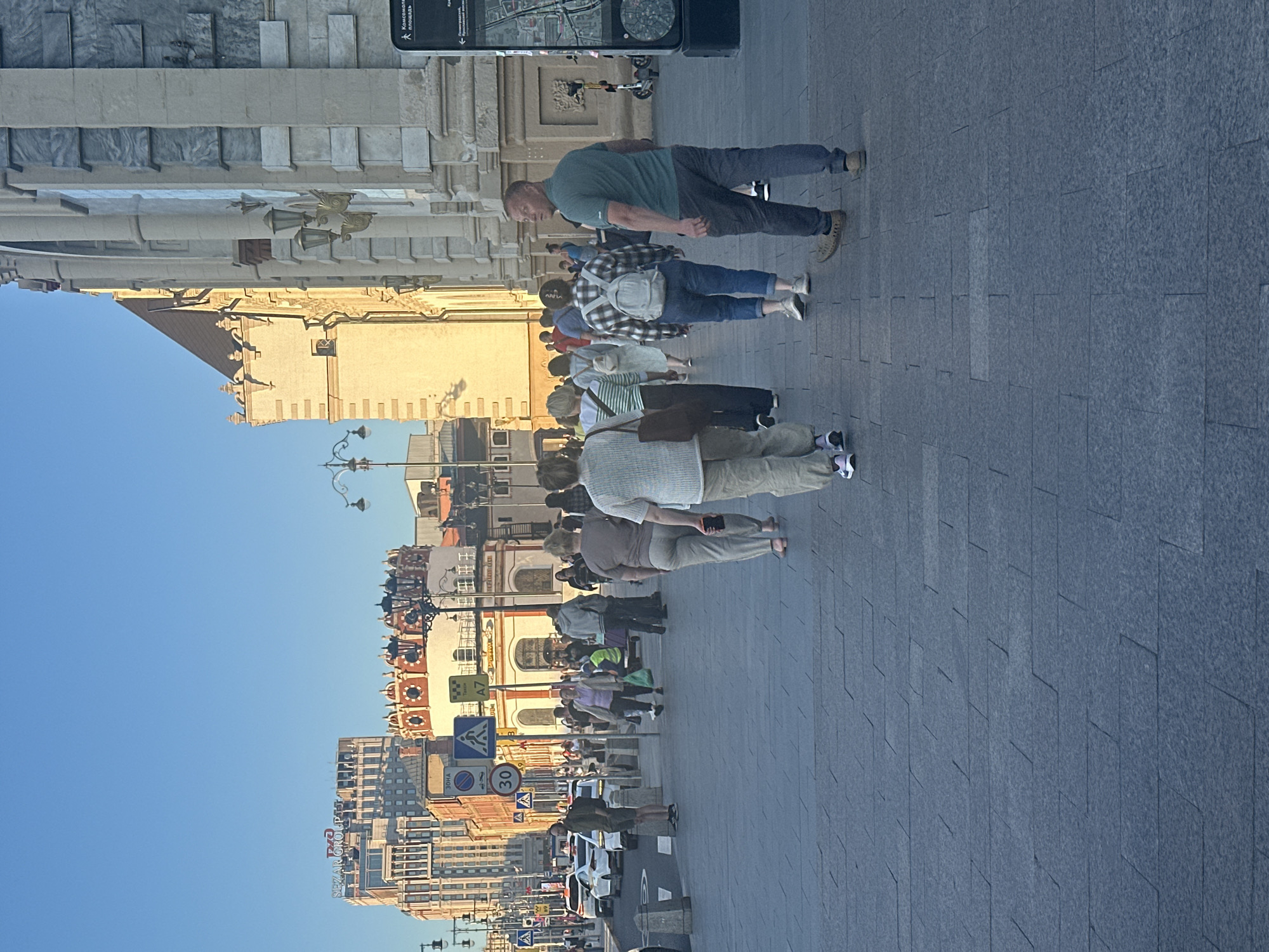 A group of people are walking on a paved sidewalk next to a street with cars. The buildings in the background appear to be old and ornate, with architectural details like columns and statues. The sky is clear and blue, suggesting it is daytime and the weather is fair. One of the people in the foreground is wearing a green t-shirt and jeans, while another is wearing a plaid shirt and carrying a backpack. There are several signs visible along the street, including traffic signs and advertisements. One sign has Cyrillic text on it, indicating a location where this alphabet is used. Another sign shows a speed limit of 30. The overall scene depicts a busy urban environment with people going about their day.