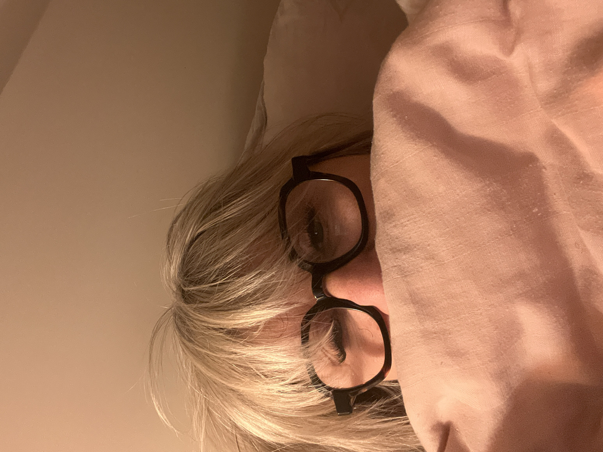 A close-up, slightly off-center shot shows a person partially obscured under a pinkish-peach fabric or blanket. The person has blonde hair and is wearing black-framed glasses. Only the person's eyes, nose, and part of their face are visible. The background appears to be a light beige or off-white wall. The image is shot indoors under warm lighting. The location of the image in Chambœuf, France, is not discernible from the visual data provided.