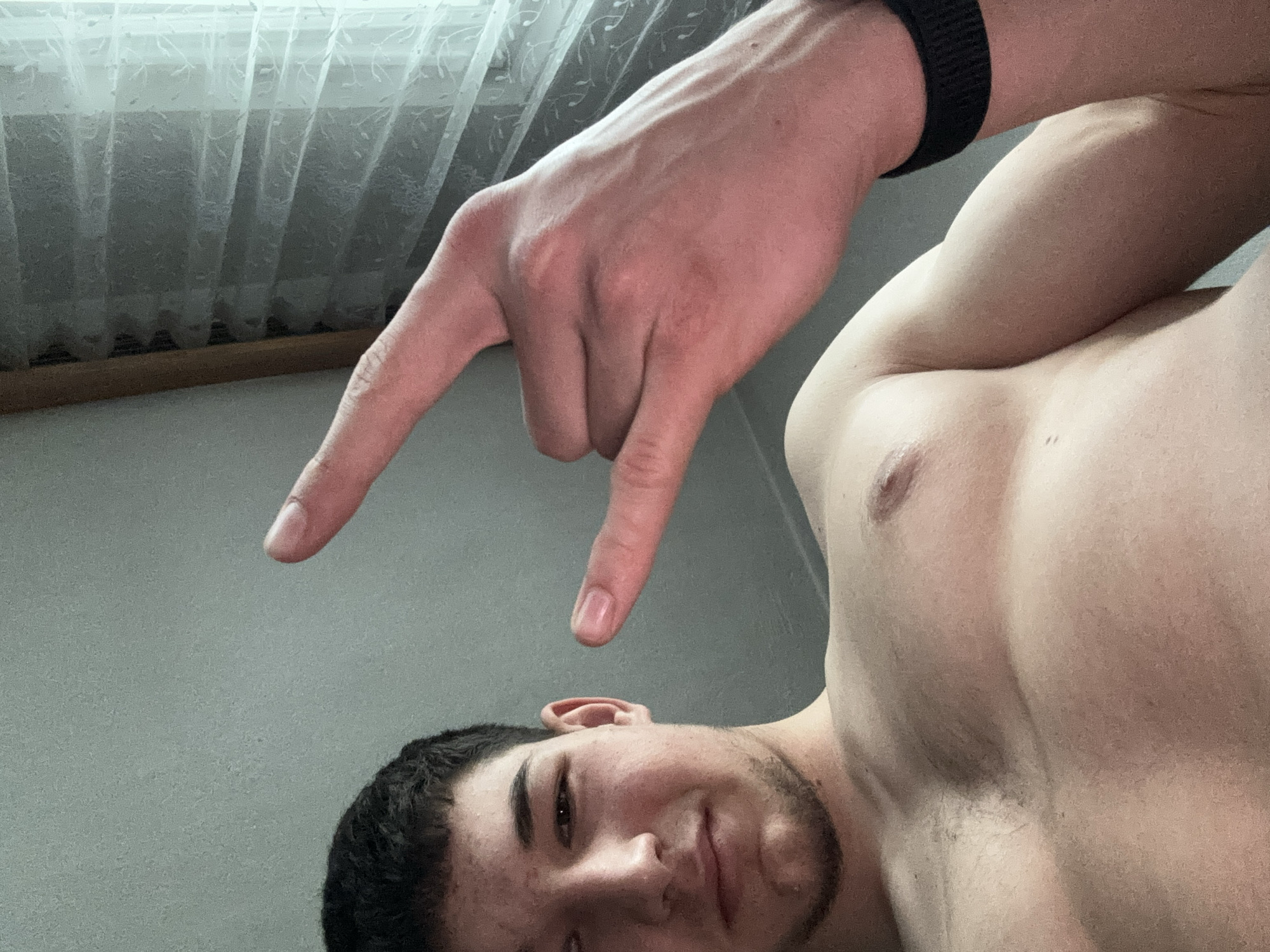 An upward-facing, rotated close-up selfie captures a man and his hand in an indoor setting in Zlatograd, Bulgaria. The primary subject is a young man, visible from his chin to just above his hairline, looking directly at the camera with a slight, friendly smile. He has short, dark hair and some stubble on his chin and jawline. To the right, part of his bare chest and shoulder are visible, suggesting he might be shirtless or wearing something low-cut.
Prominently featured above his head is a bare arm, wearing a black wristband, making the "rock on" or "devil horns" hand gesture with the index and pinky fingers extended and the middle two fingers curled down. The skin on the hand and arm shows visible veins, indicating definition.
In the background, a white, sheer curtain with a subtle patterned design covers what appears to be a window, letting in diffused daylight. A dark wooden window frame is visible along the bottom edge of the curtain. The overall lighting is soft and even. No text is visible in the image.