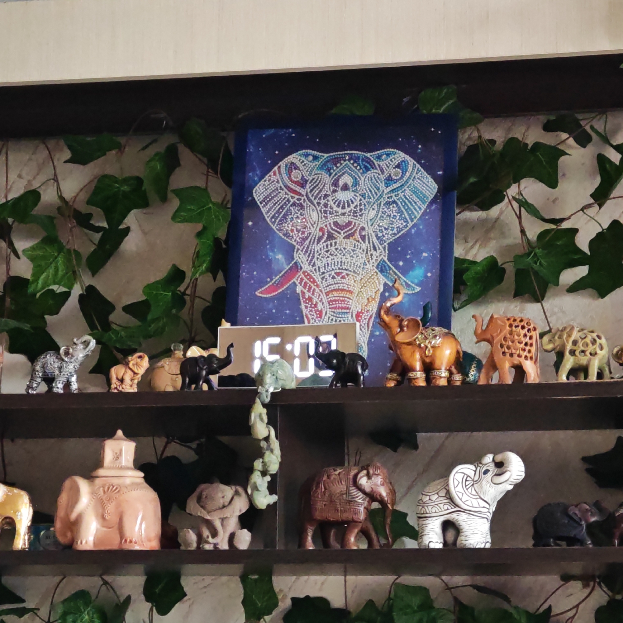 Two shelves are adorned with elephant figurines of various colors, sizes, and designs. A digital clock is located on the top shelf. Behind the figurines is a painting of an elephant and artificial ivy. The location is an interior of an unknown building in an unknown city.