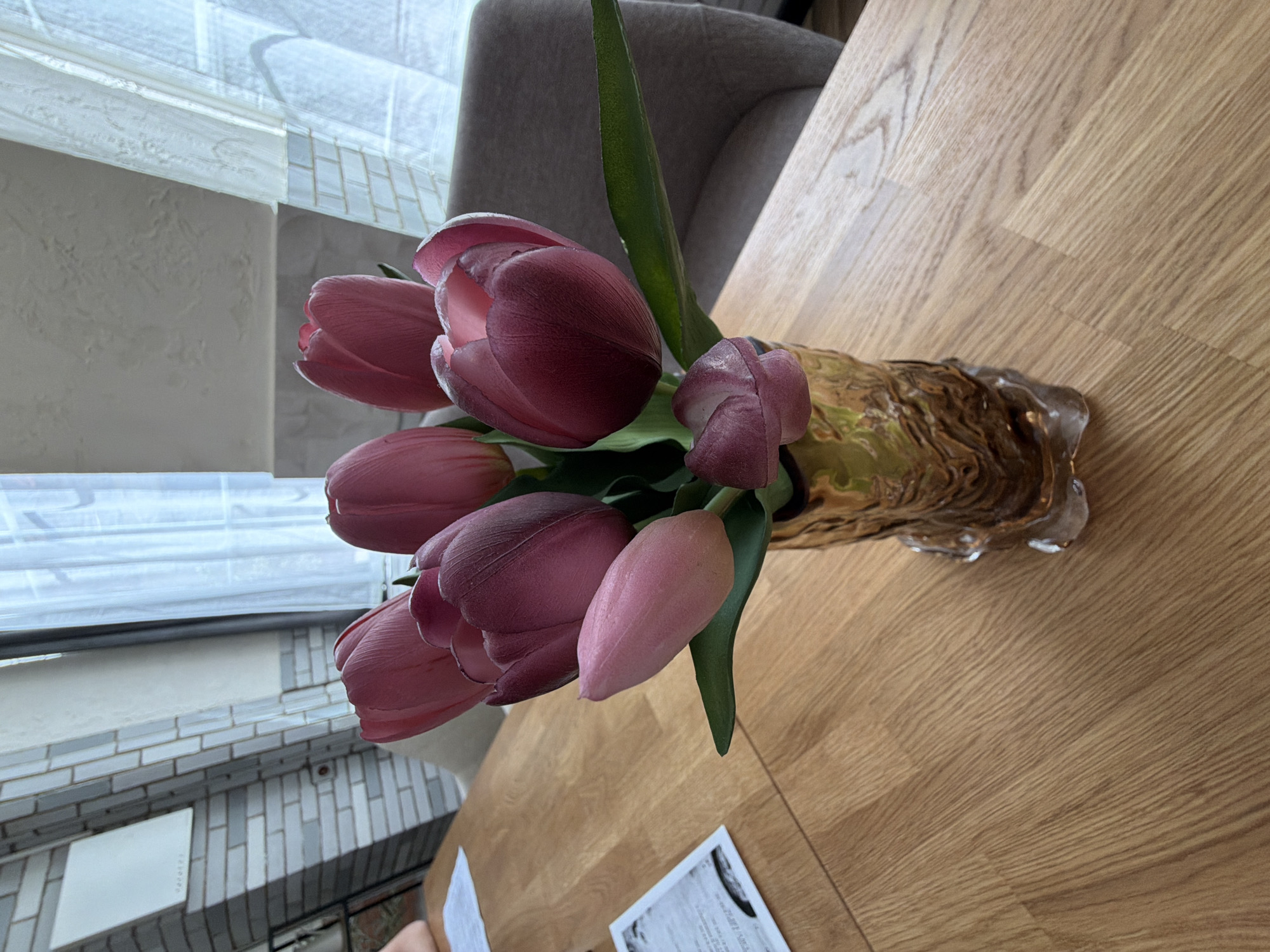 An indoor, brightly lit photograph captures a bouquet of fresh tulips arranged in a decorative vase on a light wooden table. The image, originally rotated 90 degrees counter-clockwise, depicts the tabletop extending from the bottom right to the top left.
The vase, positioned centrally, is gold-toned with a textured, possibly glass or ceramic, surface that resembles molten or organic shapes, featuring clear, ice-like details at its base. The tulips, mostly in shades of pink and deep magenta or plum, are semi-closed, revealing hints of lighter pink petals inside. Several vibrant green leaves are visible among the flower stems.
The wooden table features a distinct grain pattern, characteristic of laminate or light oak. In the top background, a grey upholstered chair or sofa back is partially visible. The left side of the frame is dominated by a large window with sheer white blinds or curtains, allowing soft, natural daylight into the room. Beyond the window, an exterior wall can be seen, characterized by light-colored, possibly white or grey, rectangular and square tiles or bricks arranged in a decorative pattern. A small, faint red circular element is present on this exterior wall.
On the table in the bottom right corner, a piece of white paper with black printed text is visible, though the content is illegible. The scene evokes a sense of calm and natural beauty, suggesting a daytime setting in a home in Silistra, Bulgaria.