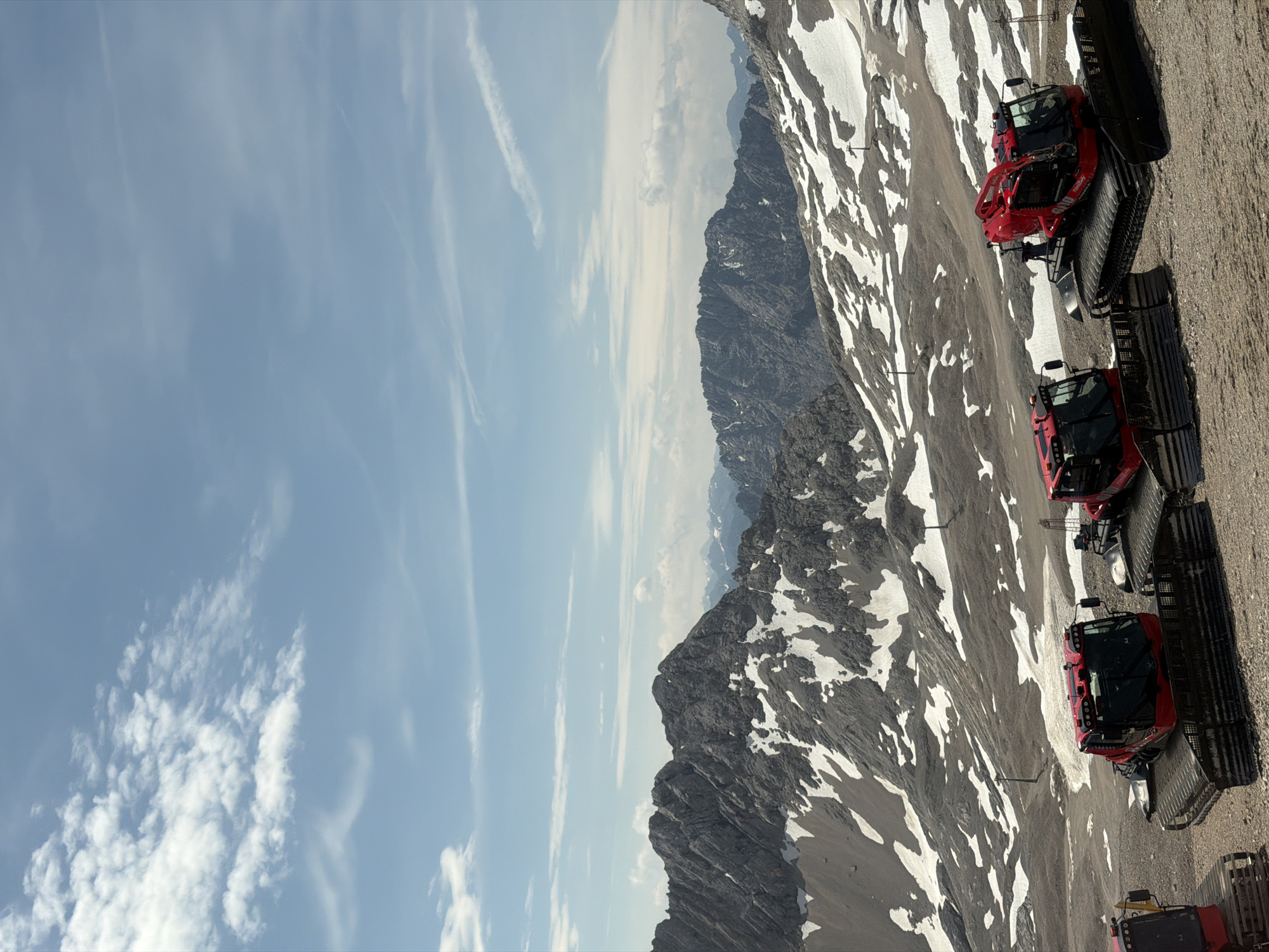 The image depicts a panoramic mountain landscape with snow patches, featuring three red snow grooming vehicles parked on a gravelly terrain. The sky above is blue with scattered white clouds. In the foreground, the snow groomers are prominently displayed. The middle ground showcases rugged, gray mountain peaks with patches of snow, suggesting a high-altitude environment. In the background, additional mountain ranges fade into the distance under the vast sky. The scene is likely situated in the Bavarian Alps, near Garmisch-Partenkirchen, Germany, a region known for its mountainous terrain and winter sports facilities. No people are visible in the image. The overall atmosphere is serene and suggests a location that experiences significant snowfall and is prepared for winter activities.