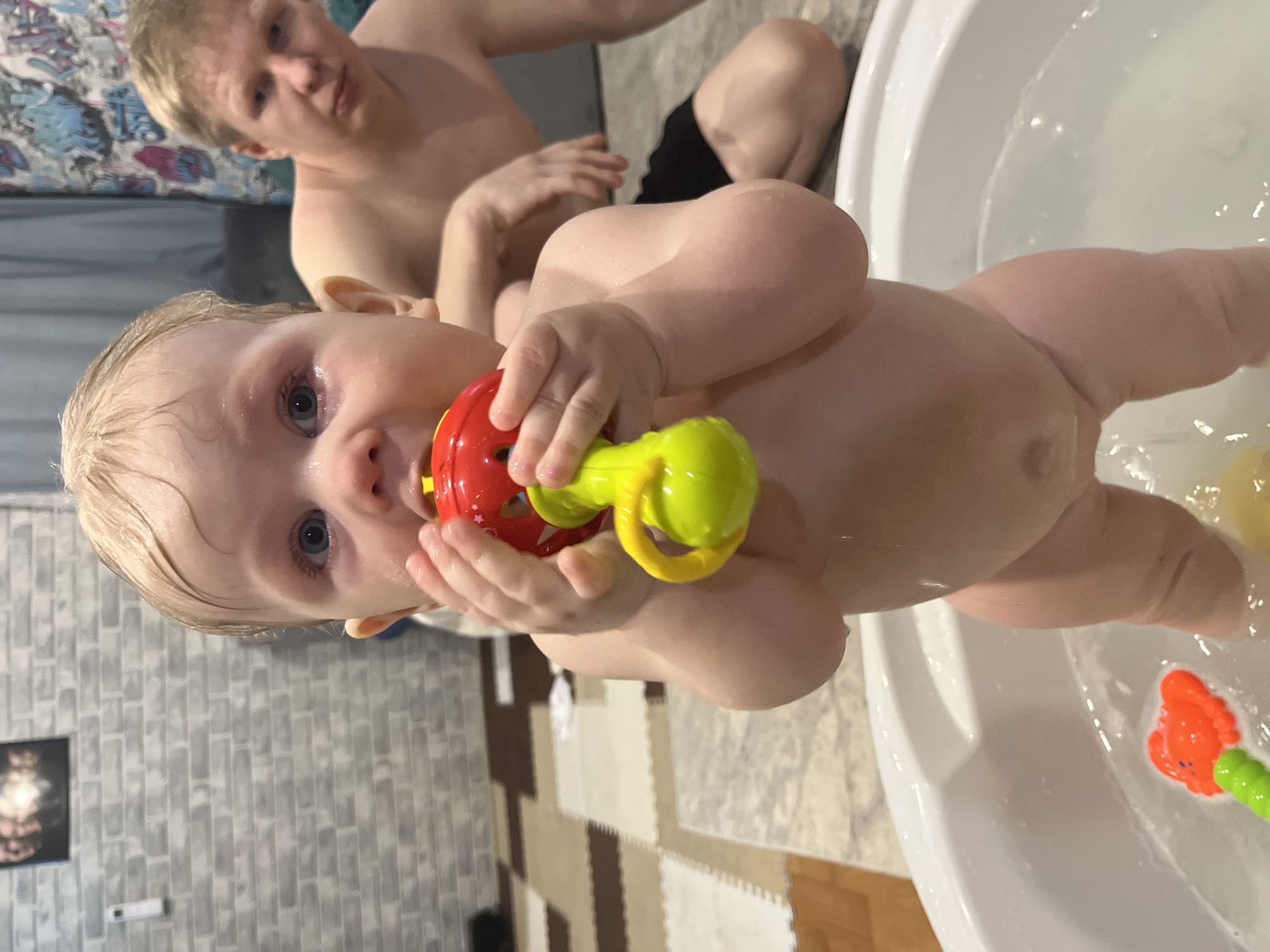 A young child is taking a bath in a white plastic tub. The child has wet blonde hair, blue eyes, and is holding a red and green plastic toy. An adult is in the background wearing no shirt and black shorts. The location appears to be indoors, with a gray brick-like wall and a puzzle mat on the floor. The specific location in Vesyoloye, Russia, cannot be determined from the image.