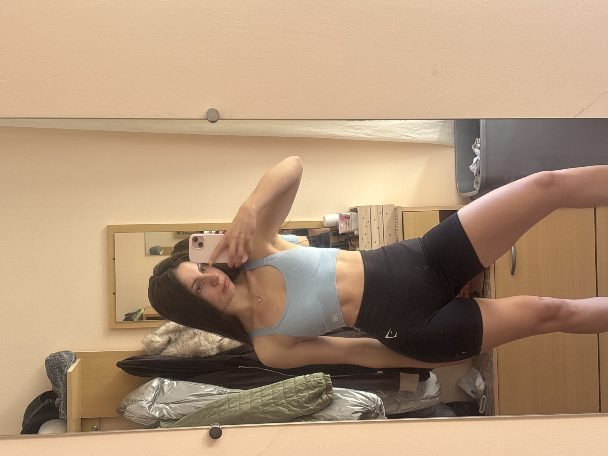 A young woman is seen in a mirror selfie. She wears a light blue sports bra and black shorts. Her hair is dark and long. She holds a pink iPhone in her hand. Reflected behind her is a room with a bed covered with clothes, including a silver jacket and a green quilted jacket. A wooden cabinet and a grey container are also visible. The exact location within Varna, Bulgaria is indeterminable from the image.