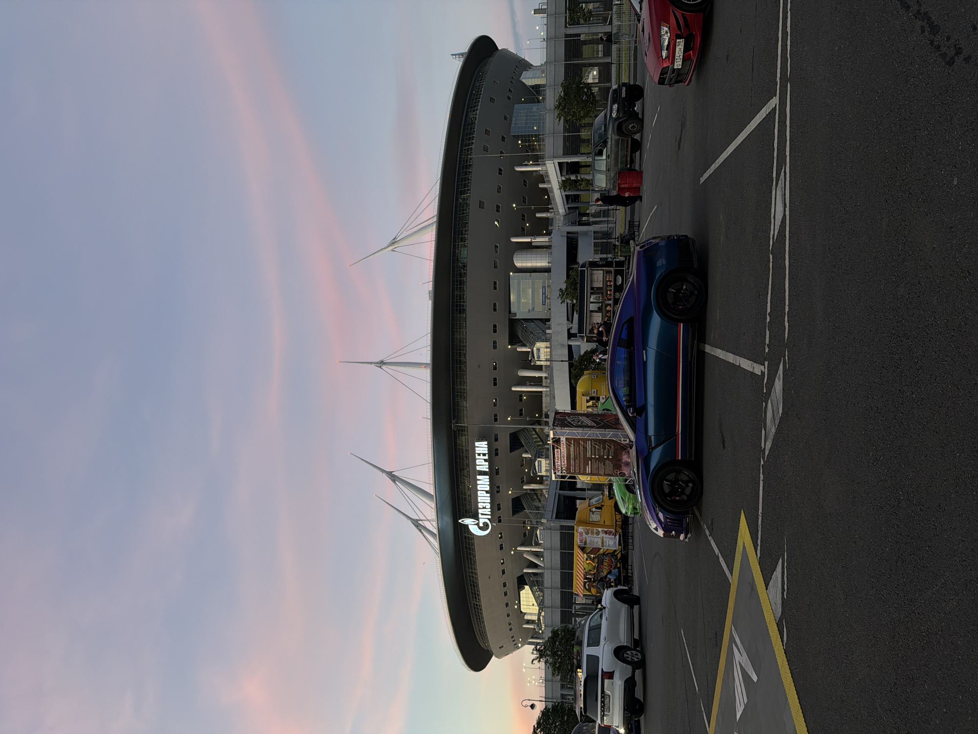 The image depicts Gazprom Arena in Saint Petersburg, Russia, under a pastel-colored twilight sky. Several cars are parked in a lot in front of the stadium, including a blue Nissan GT-R, a white SUV, a dark green SUV, and a red Mitsubishi Lancer. Food trucks are also visible, suggesting an event or gathering. The stadium itself is a prominent, modern structure with a distinctive roof design. The overall scene is one of urban activity at dusk, with a focus on both the architectural landmark and the vehicles.